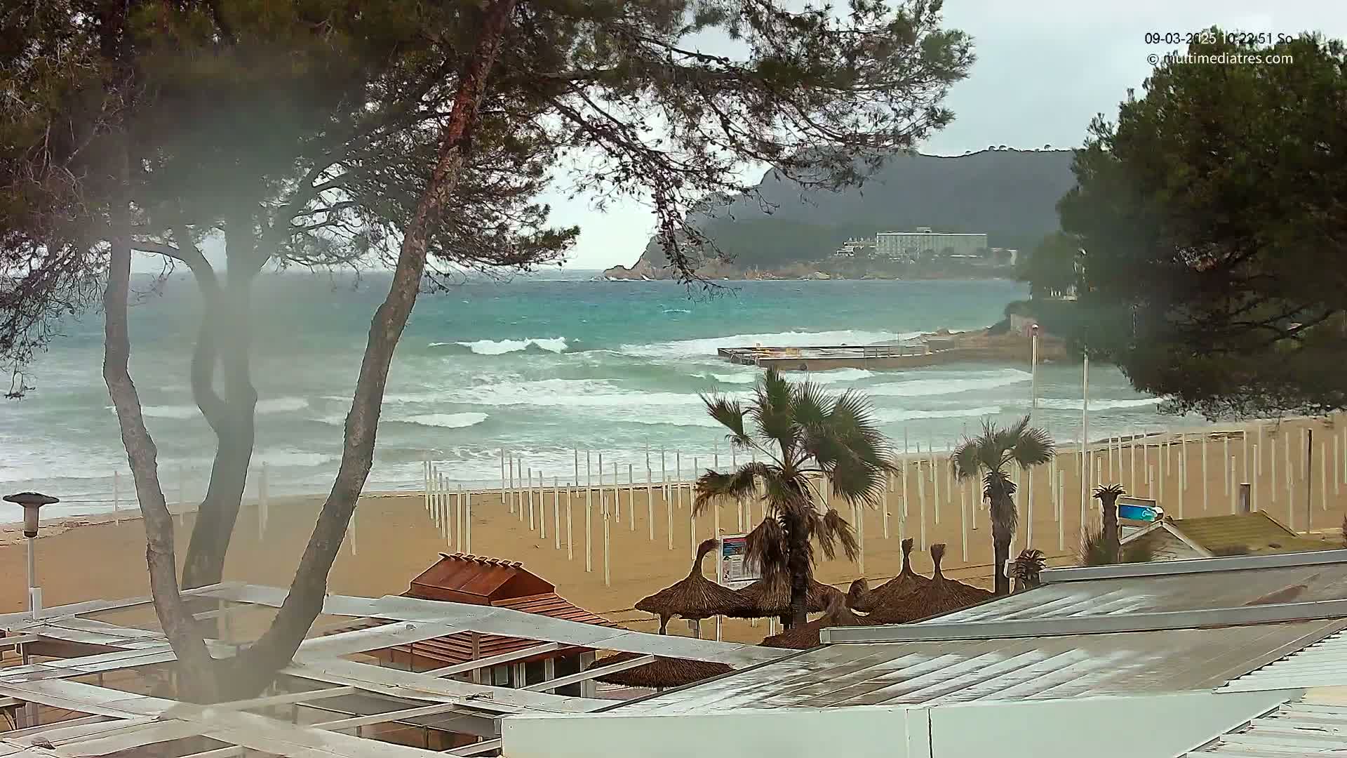 A beach with choppy waves under an overcast sky, seen from behind beachside structures and trees.