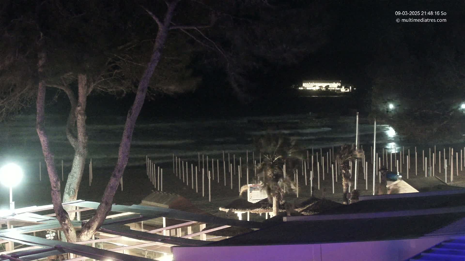 A nighttime beach scene shows a dark, choppy ocean with a distant, illuminated building, a row of poles on the beach, and a tree in the foreground under a partly-lit outdoor structure.