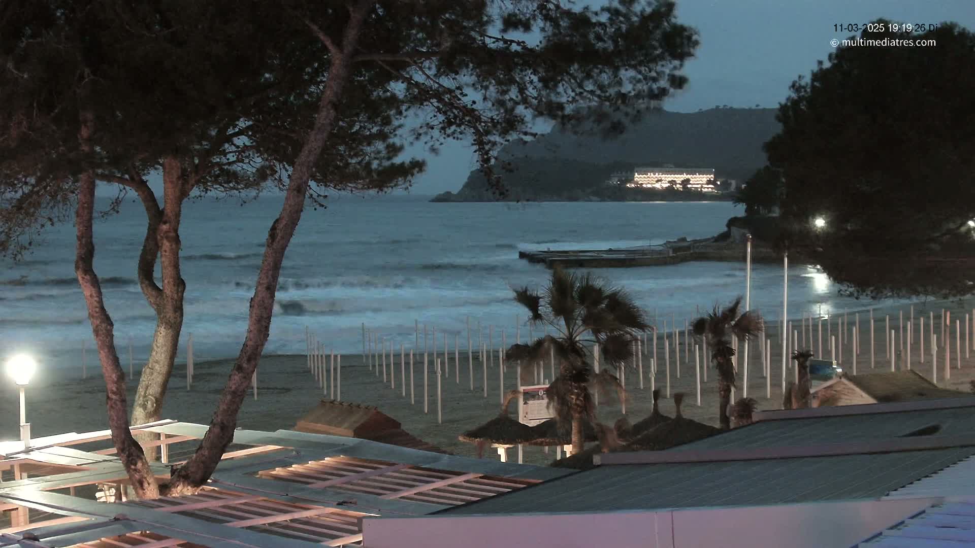 A nighttime beach scene with moderately rough waves under a dark, cloudy sky, showing a sandy beach with palm trees and beach structures.