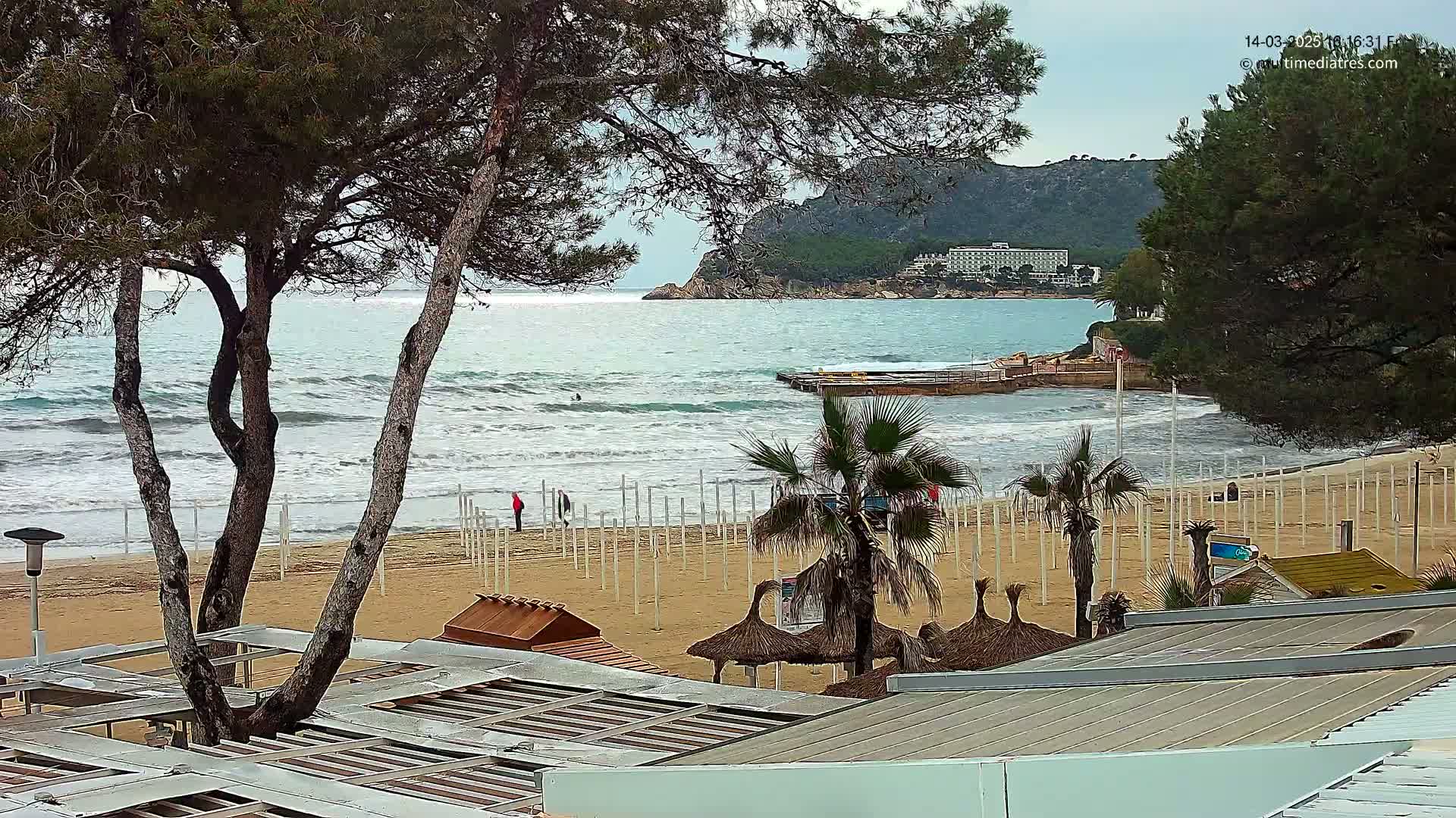 A sandy beach with moderate waves, palm trees, and beach structures is visible, with a hotel and a rocky hill in the background under a partly cloudy sky.