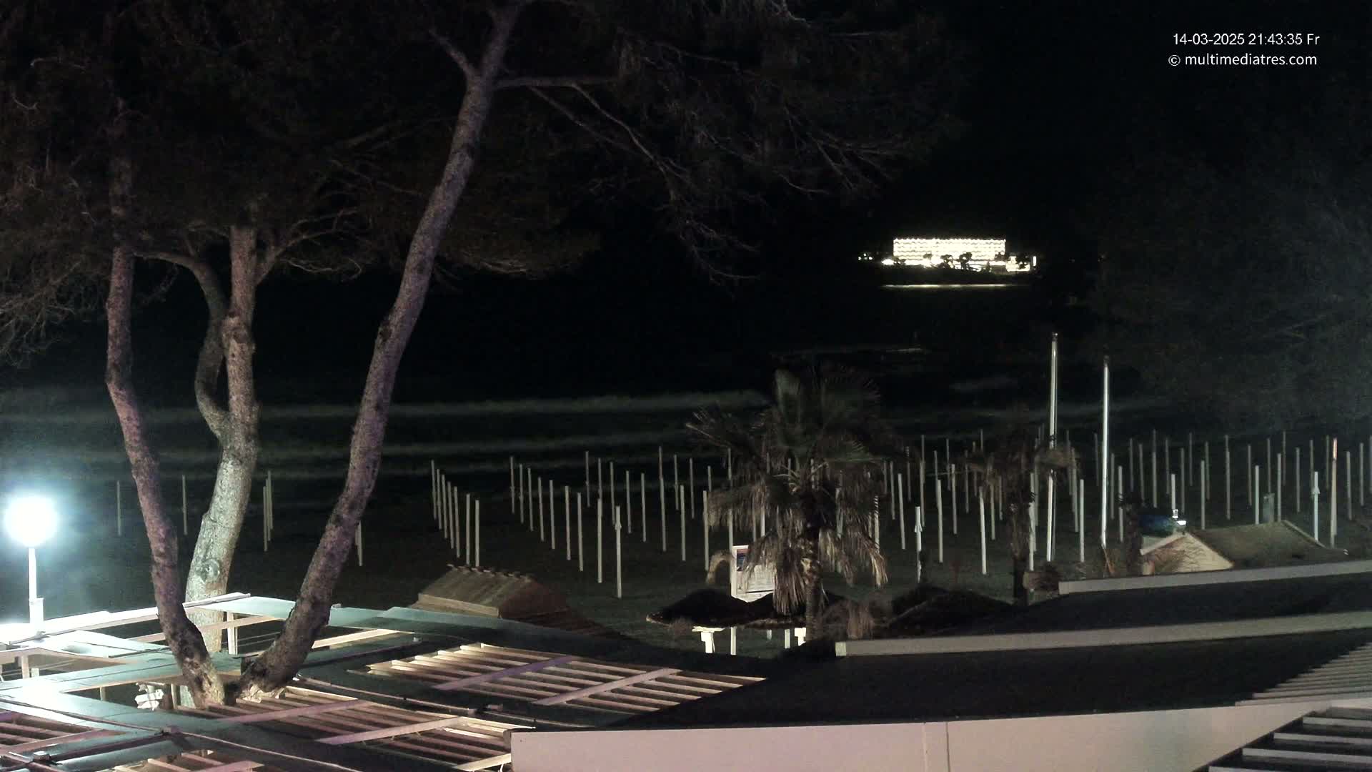 A nighttime beach scene shows dark trees, a sandy shore with numerous upright posts, a dimly lit building in the distance, and calm waves lapping the shore.