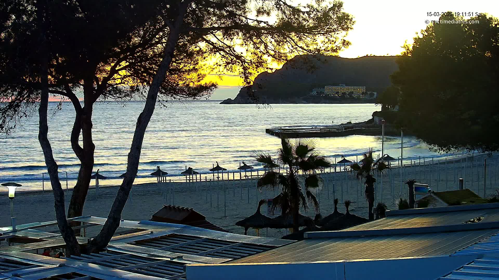 A sandy beach with palm trees and closed umbrellas is visible at sunset, overlooking a calm sea with small waves lapping the shore, under a clear sky.