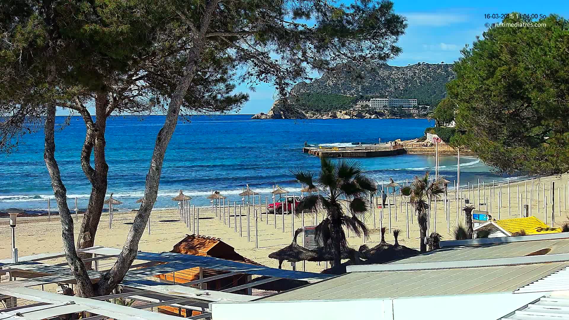 A sandy beach with rows of closed umbrellas, palm trees, and a small building is visible under a clear blue sky, overlooking a calm blue ocean and a rocky coastline.
