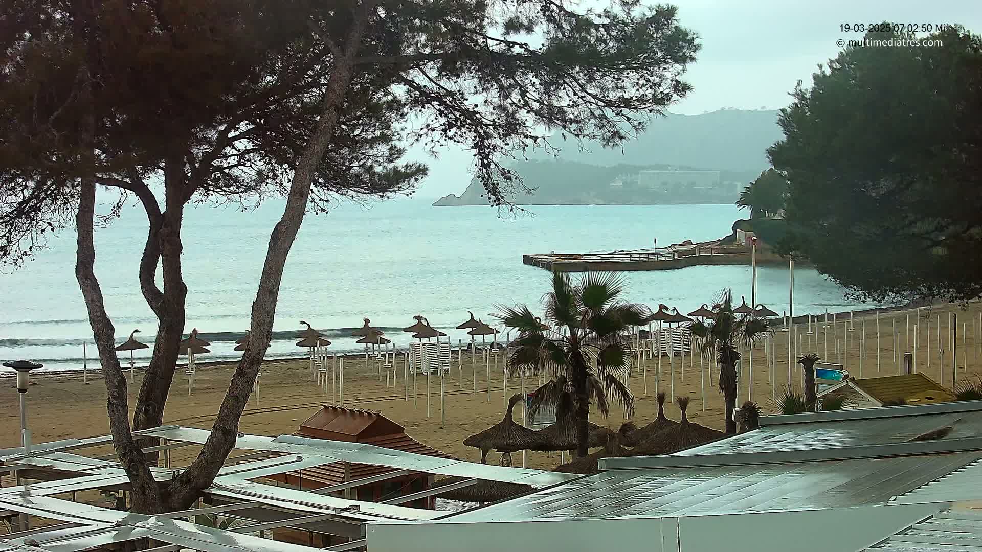 A rainy day at a beach, showing sandy shores with closed umbrellas, palm trees, and a calm sea under a cloudy sky.