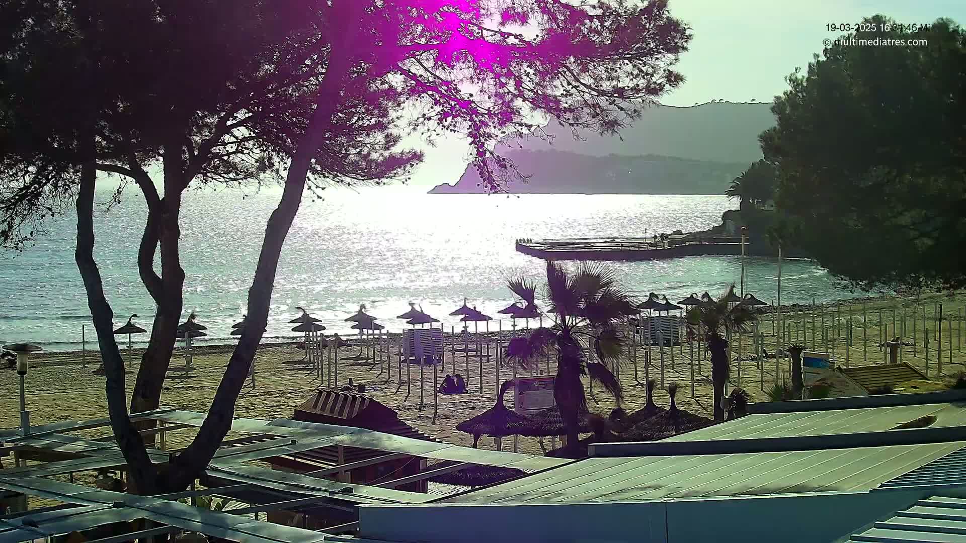A sandy beach with numerous closed umbrellas sits under a partly sunny sky beside a calm, sparkling sea, backed by trees and hills.