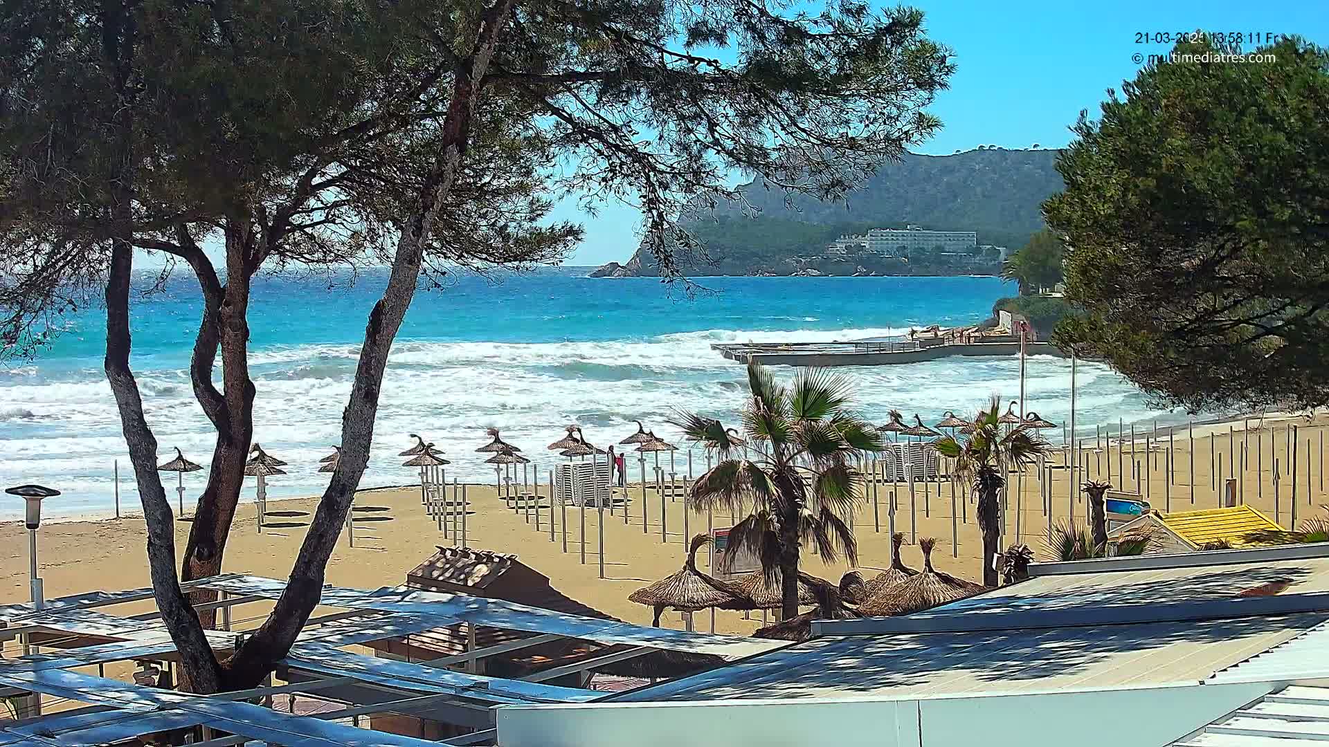 A sandy beach with numerous closed umbrellas and palm trees is partially visible under a sunny sky, overlooking a turquoise ocean with moderate waves crashing on the shore near a rocky mountain.