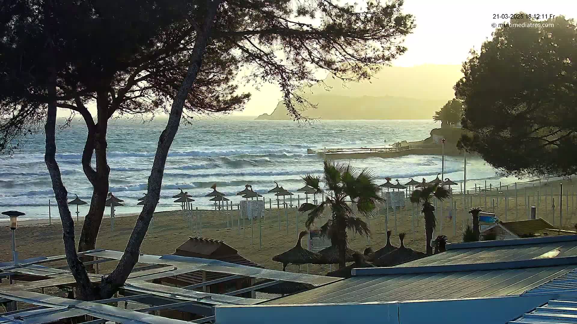 A sandy beach with rows of closed umbrellas and palm trees is viewed from behind a structure, overlooking a wavy ocean under a partly cloudy sunset sky.