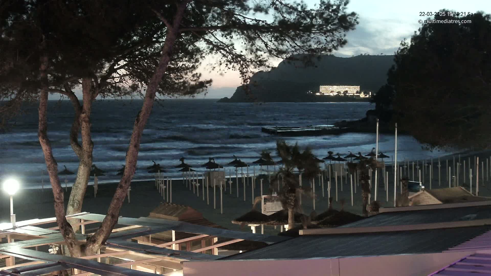 A dark beach at twilight with choppy waves, numerous closed beach umbrellas, and a distant, illuminated building on a hillside.