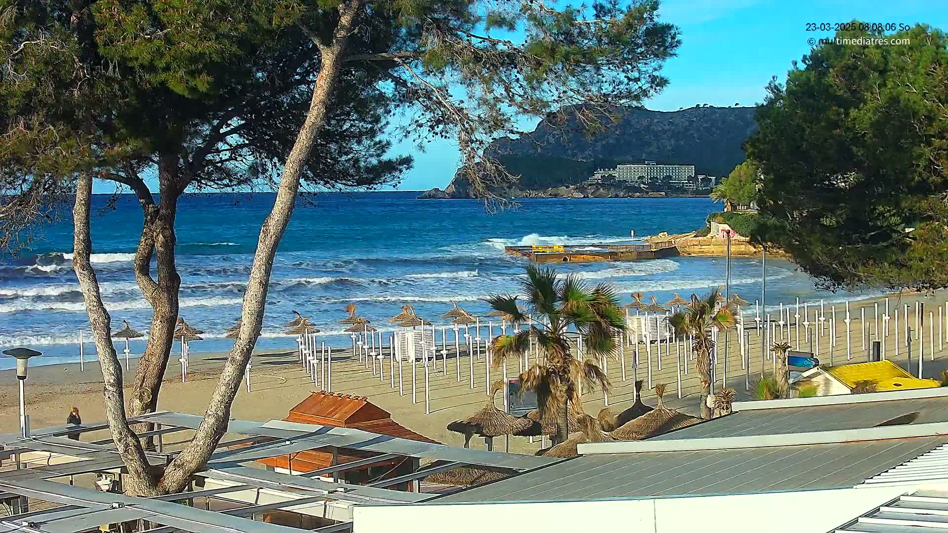 A sandy beach with rows of closed umbrellas and palm trees overlooks a wavy turquoise ocean, with a rocky hillside and distant buildings visible in the background under a clear sky.