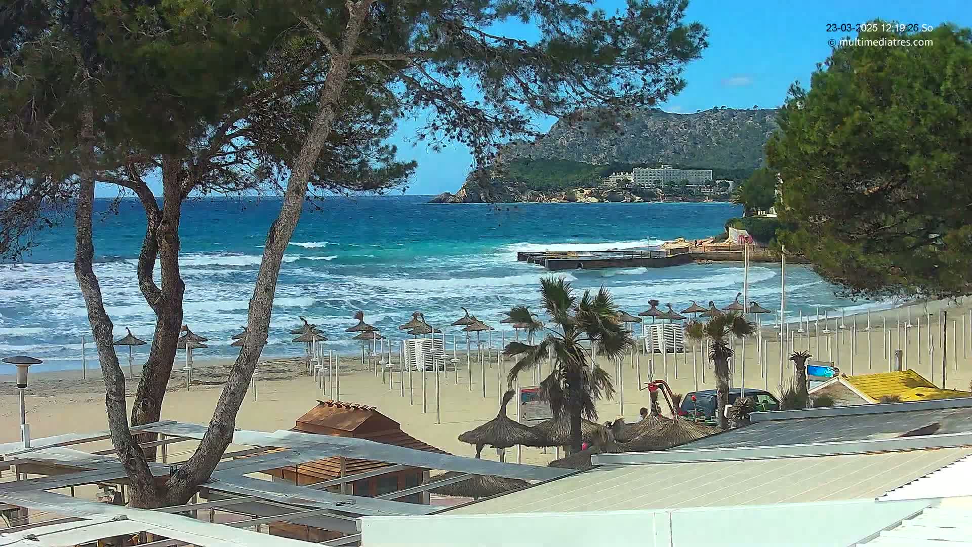 A sandy beach with numerous closed umbrellas, palm trees, and a small pier is seen next to a turquoise sea with moderate waves under a sunny sky, backed by a rocky hill with buildings.