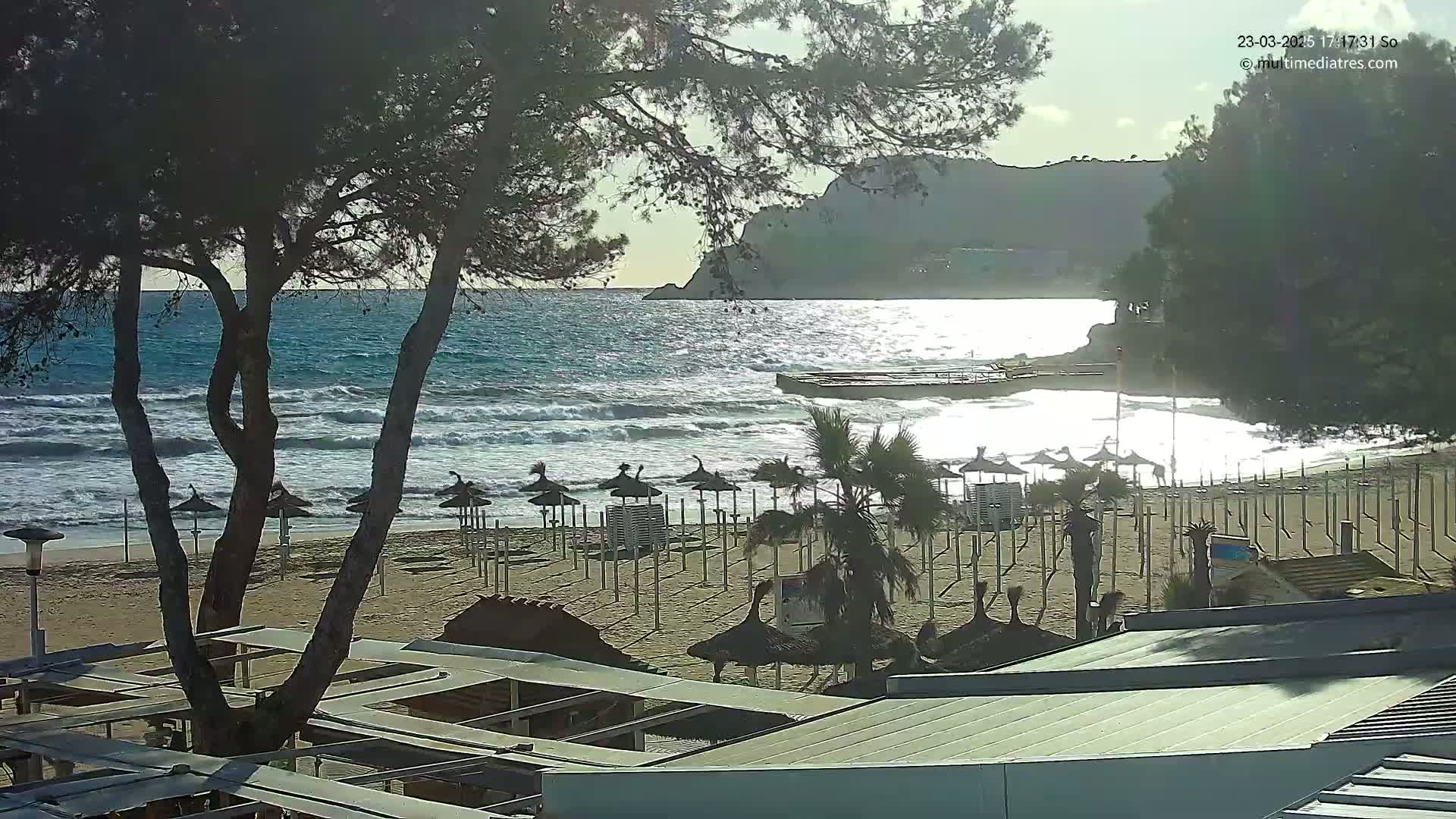 A sandy beach with numerous closed umbrellas is partially visible behind dark trees, overlooking a wavy ocean with a distant rocky shoreline under a mostly sunny sky.
