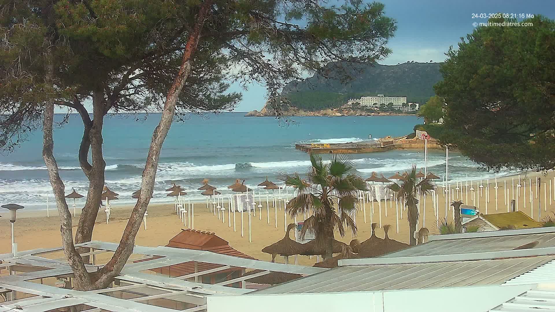 A sandy beach with numerous closed beach umbrellas under a partly cloudy sky, bordered by trees and overlooking a calm, wavy ocean with a distant shoreline and hills.