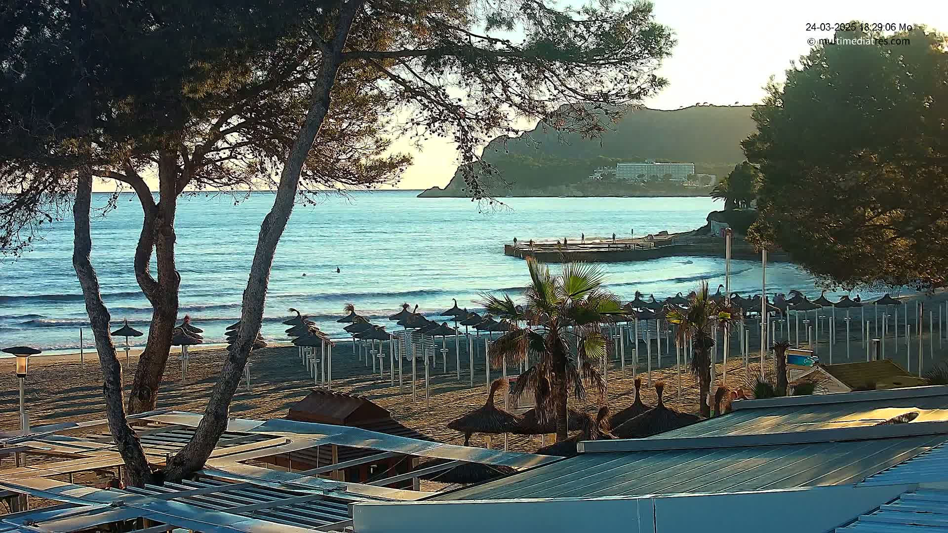 A sandy beach with numerous closed umbrellas stretches along a calm, blue ocean under a mostly clear sky, with a rocky hill visible in the distance.