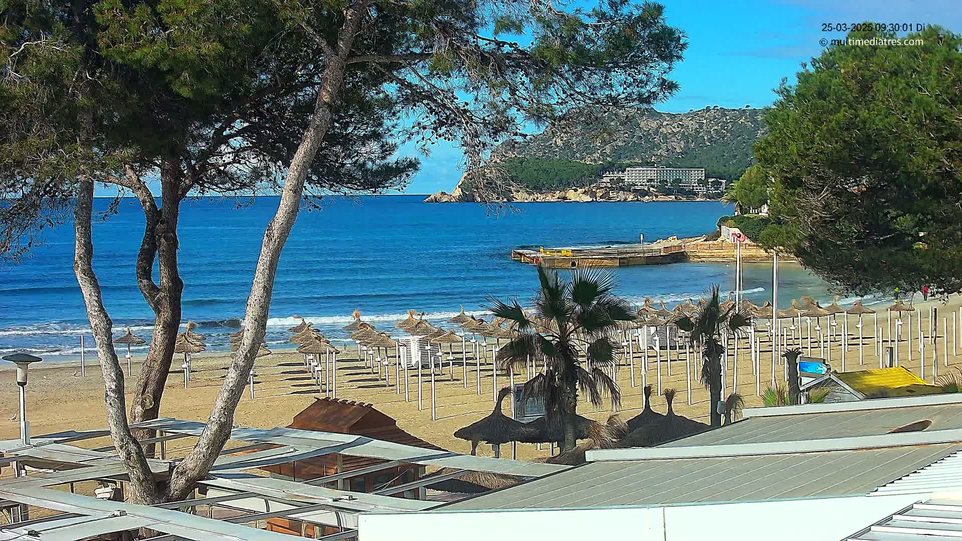 A sandy beach with numerous closed umbrellas is visible under a clear sky, overlooking a calm blue ocean and distant hills.