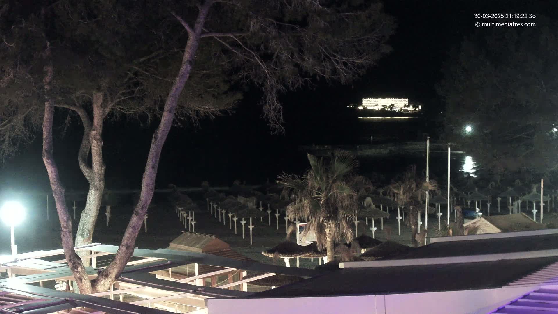 At night, a beach with numerous closed umbrellas is visible beneath a dark sky, with a brightly lit building in the distance.