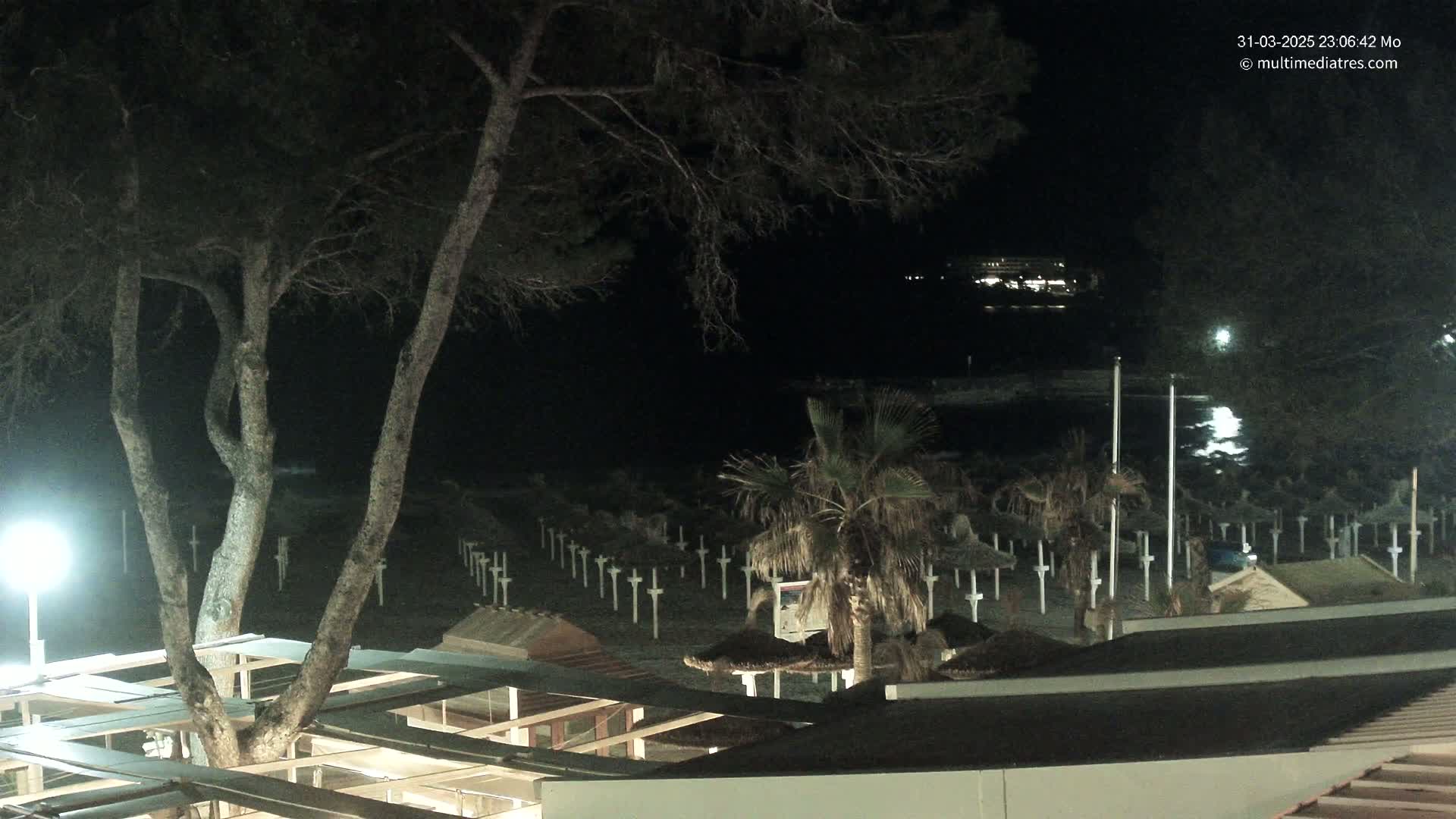 At night, a dark beach with numerous closed beach umbrellas is visible under a mostly clear sky, illuminated by artificial lights.