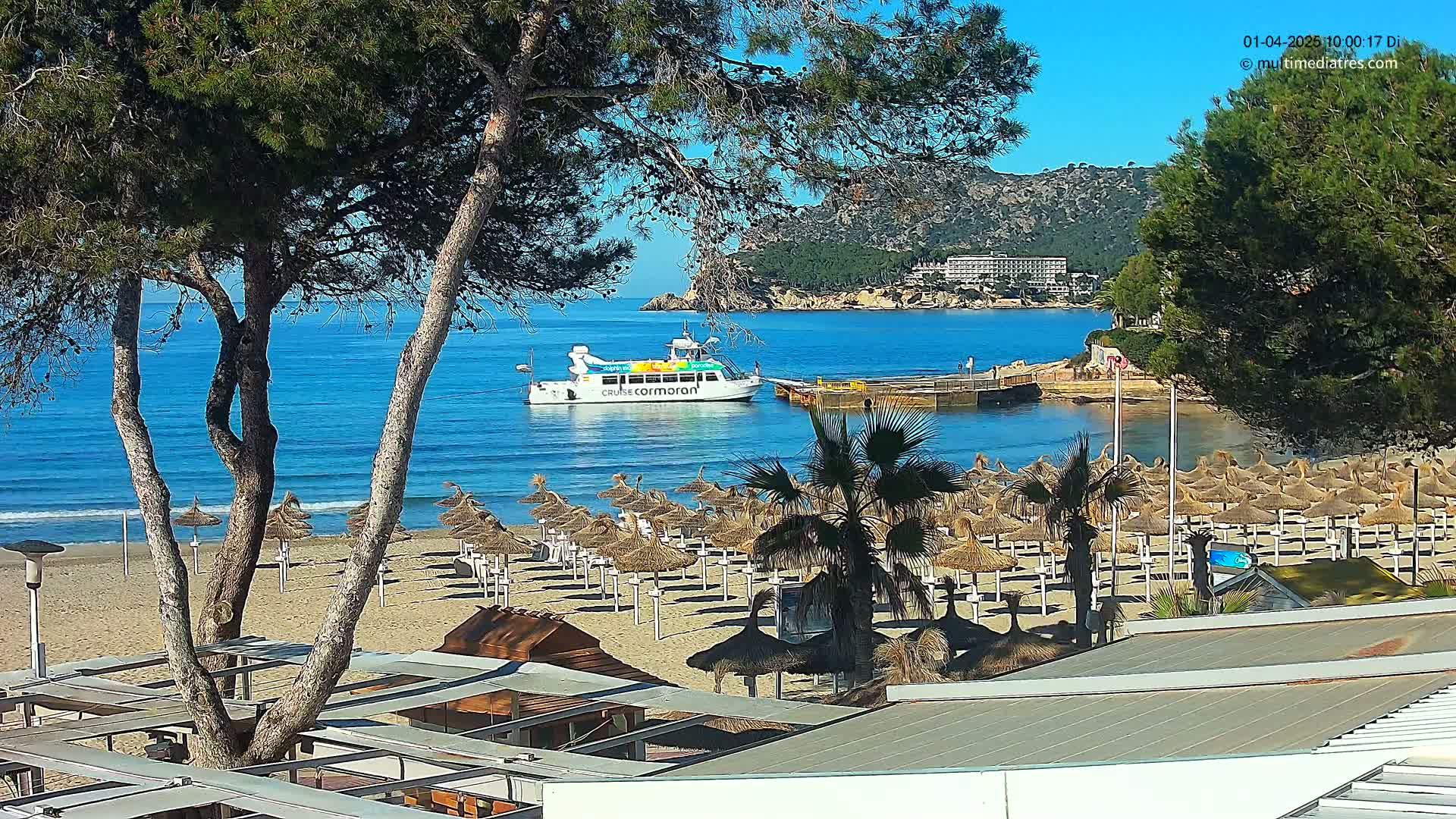 A sandy beach with numerous straw umbrellas is seen from above, with a boat in a calm, blue ocean and hills in the background under a clear, sunny sky.