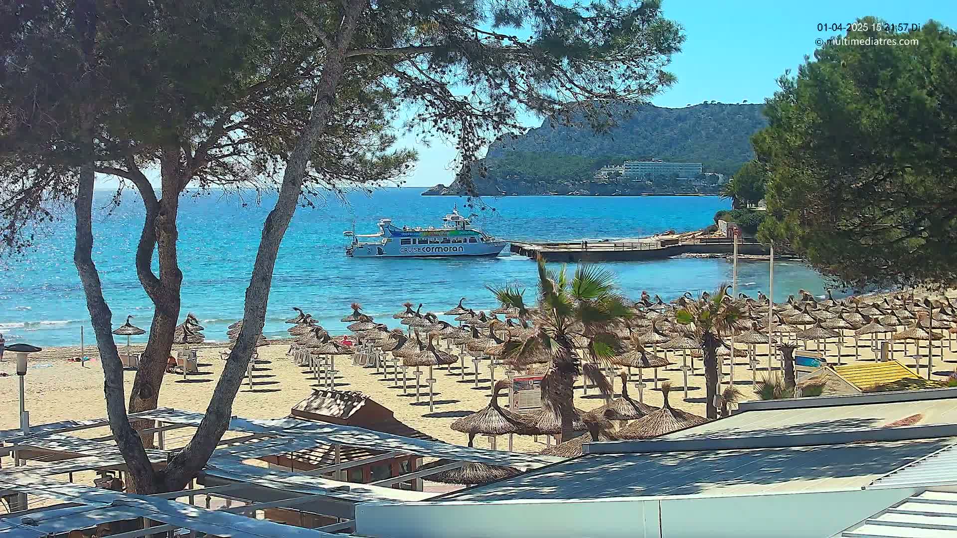 A sandy beach with numerous thatched umbrellas, palm trees, and a boat in the turquoise water under a sunny sky, backed by a mountainous shoreline.