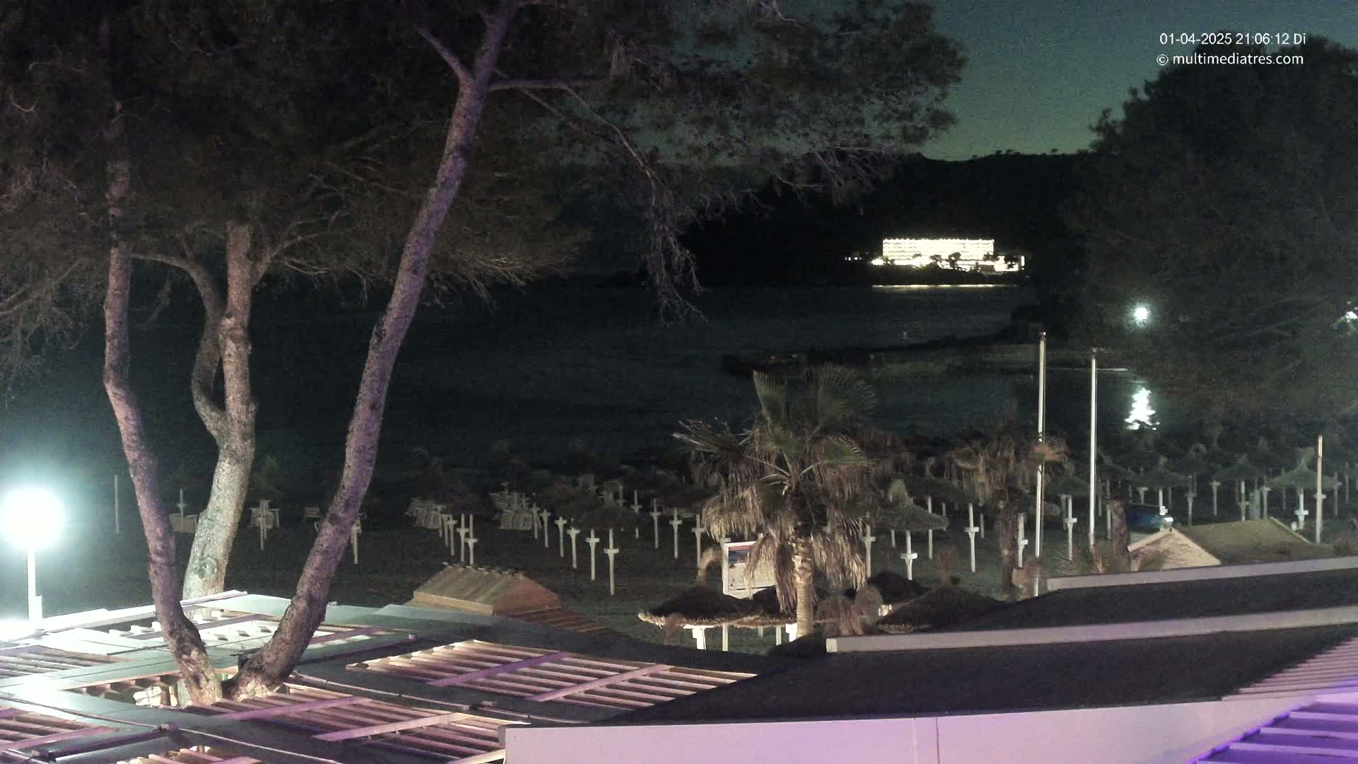 A nighttime view of a beach with numerous closed umbrellas and a building with lights visible across the water under a dark sky.