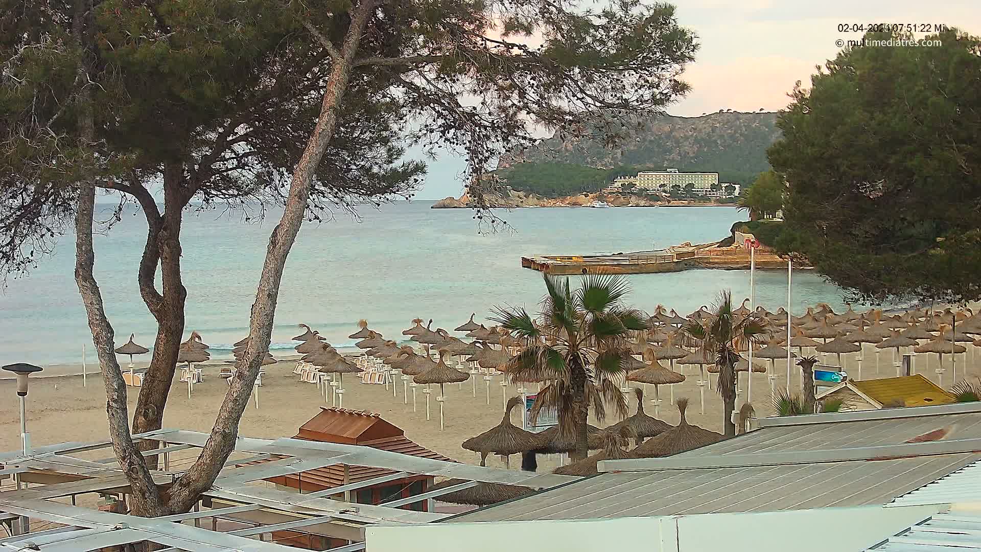 A sandy beach with numerous straw umbrellas and palm trees overlooks a calm, light-blue ocean and a distant hotel nestled against a hillside under an overcast sky.
