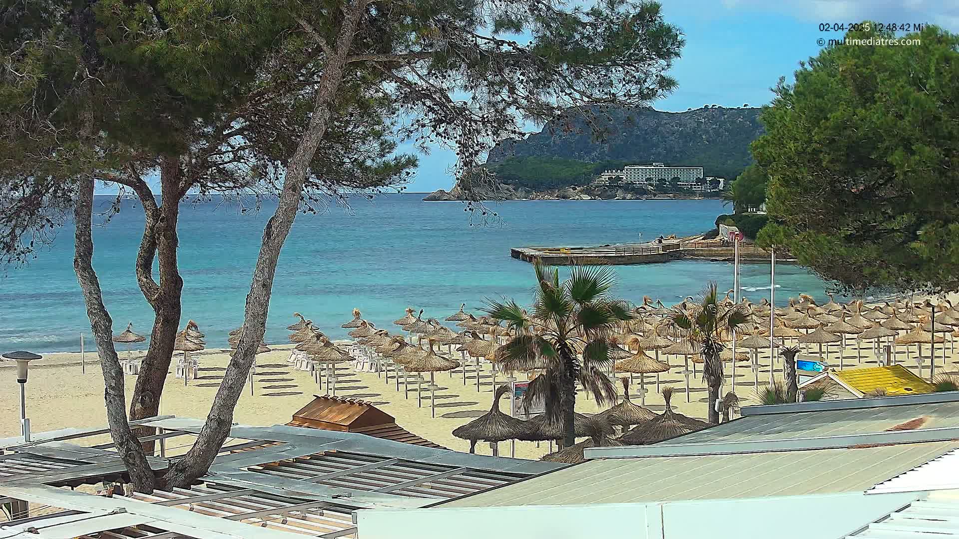 A sandy beach with numerous straw umbrellas stretches along a calm, turquoise ocean under a sunny sky, backed by a rocky hillside and trees.