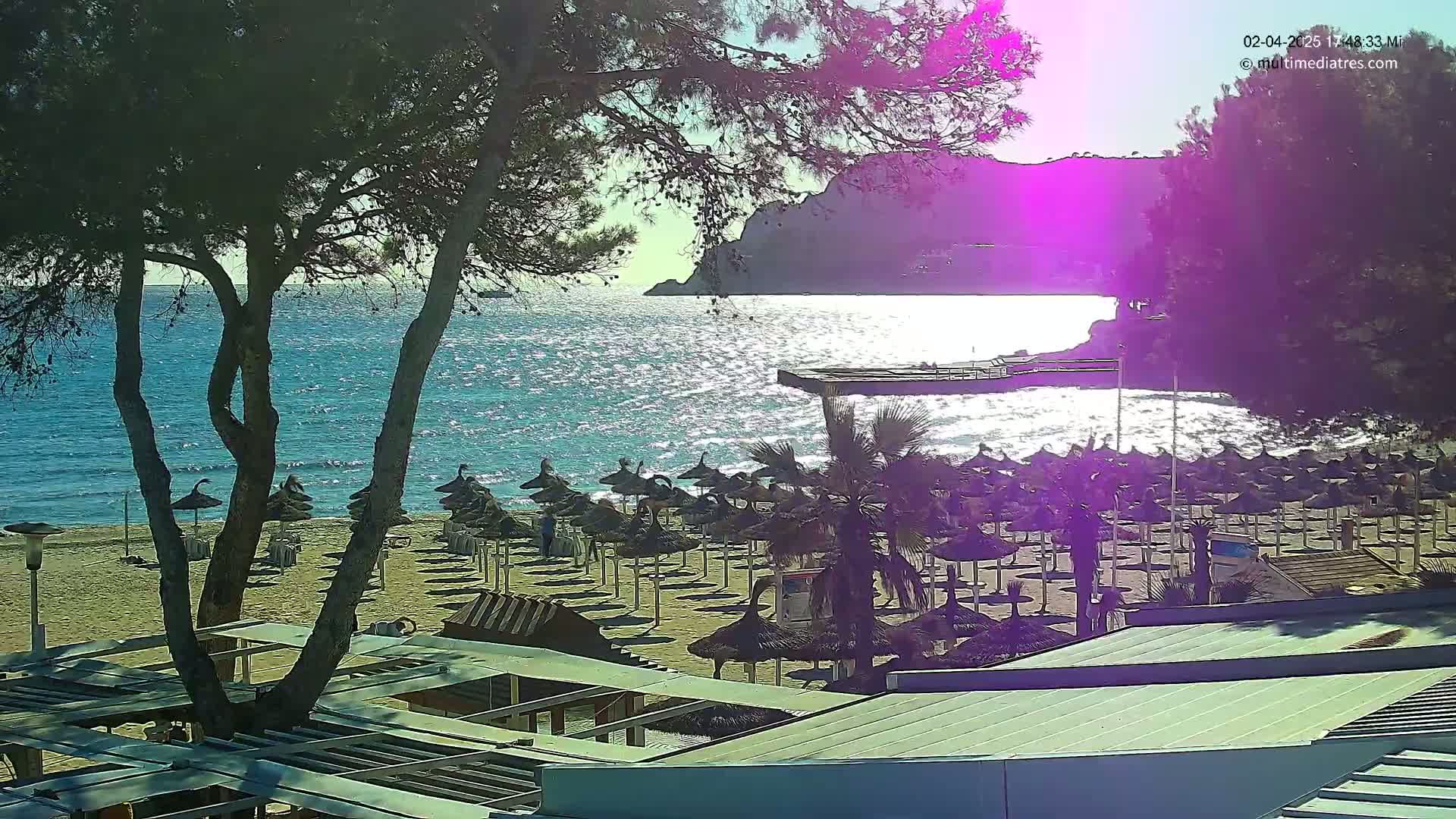 A sandy beach with numerous umbrellas and lounge chairs is visible beneath trees, overlooking a sparkling blue ocean and a distant rocky shore under a bright, sunny sky.