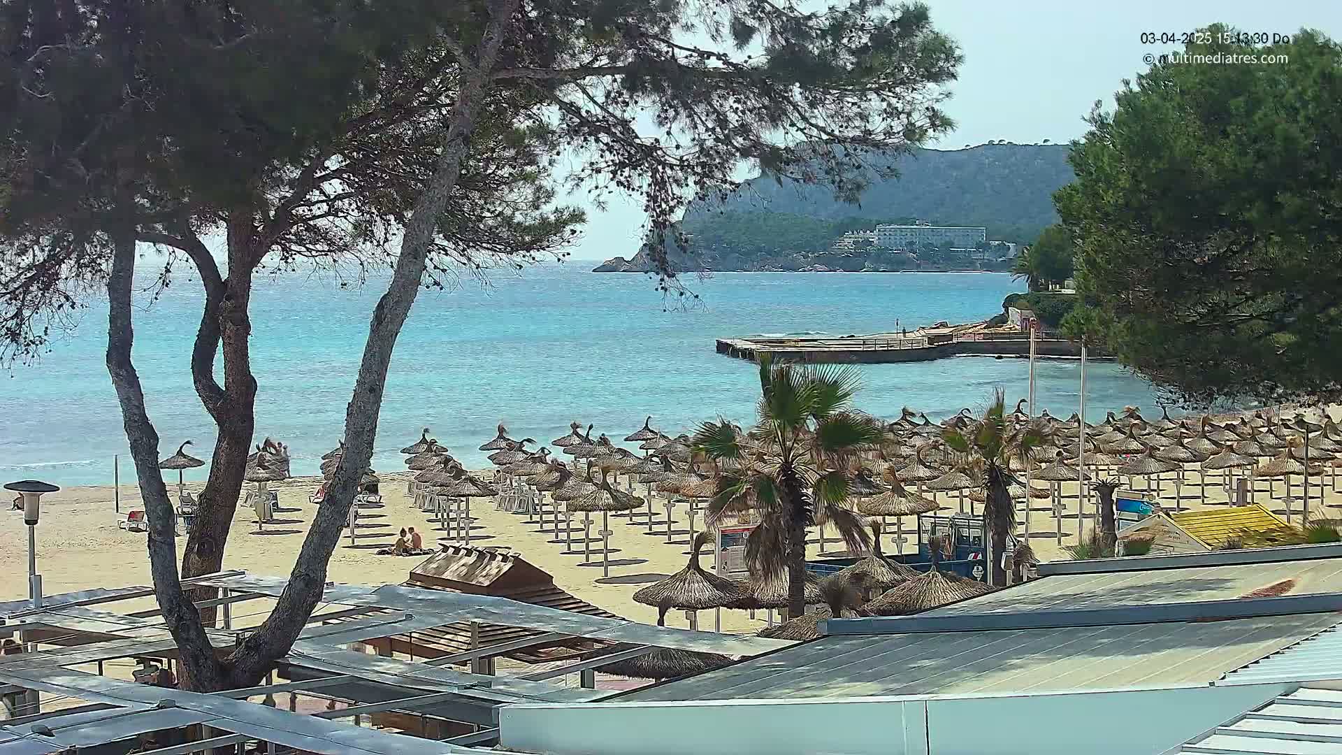 A sandy beach with numerous thatched umbrellas, palm trees, and a calm, turquoise ocean is seen under a sunny sky, with a small pier visible in the distance.