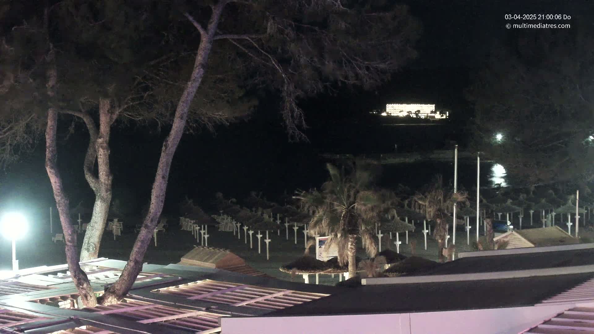 At night, a beach with closed umbrellas is seen from a rooftop vantage point, with trees and a brightly lit building visible in the distance across calm water.