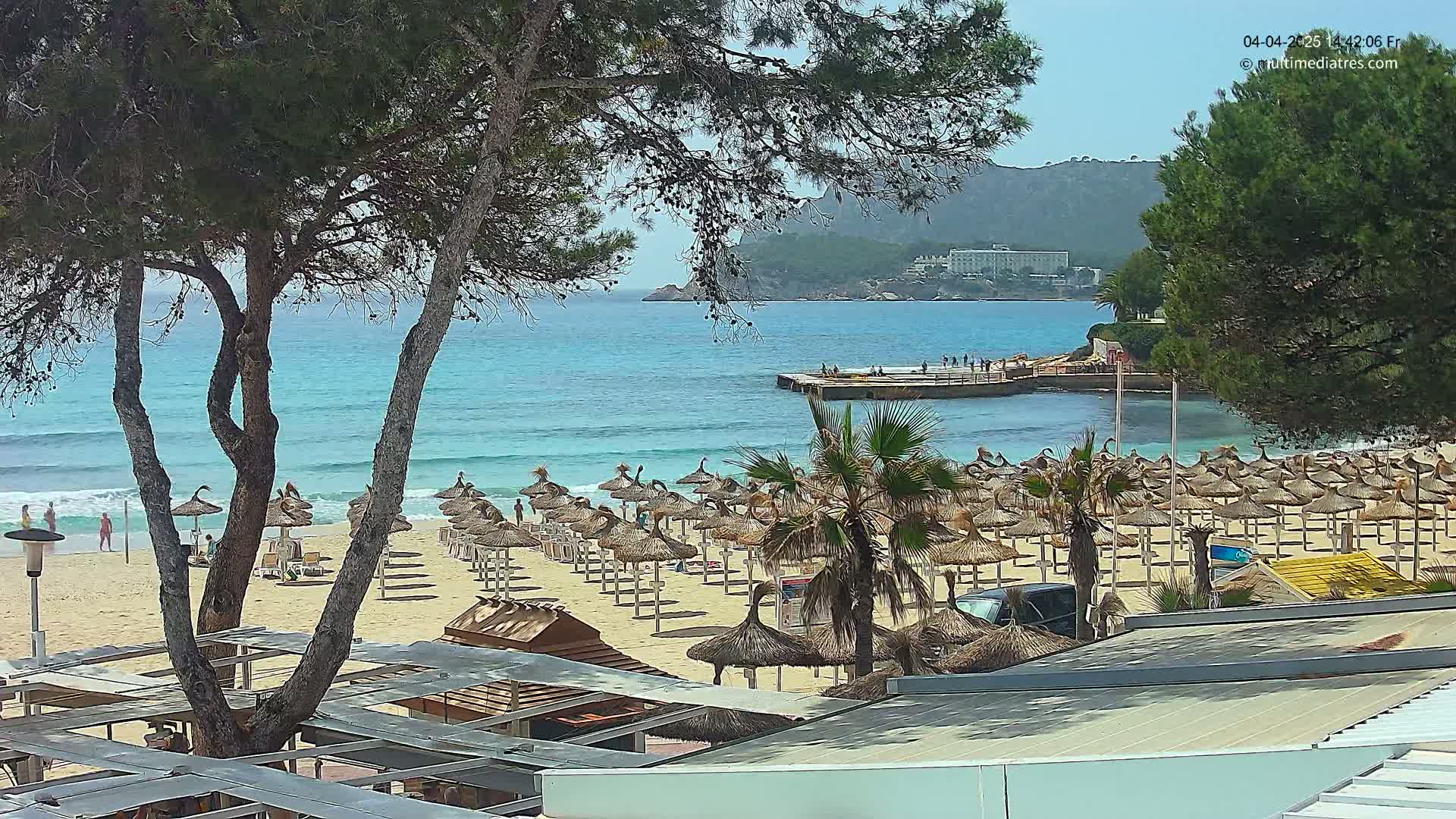 A sandy beach with numerous thatched umbrellas, calm turquoise water, and a distant rocky hillside under a sunny sky is viewed from behind a line of trees.