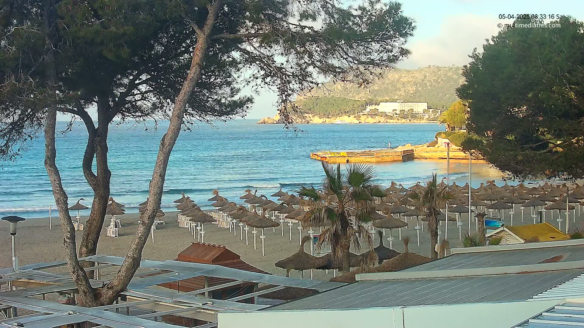 A sandy beach with numerous straw umbrellas is visible, alongside calm, blue water lapping the shore under a mostly clear sky, with hills and buildings in the background.