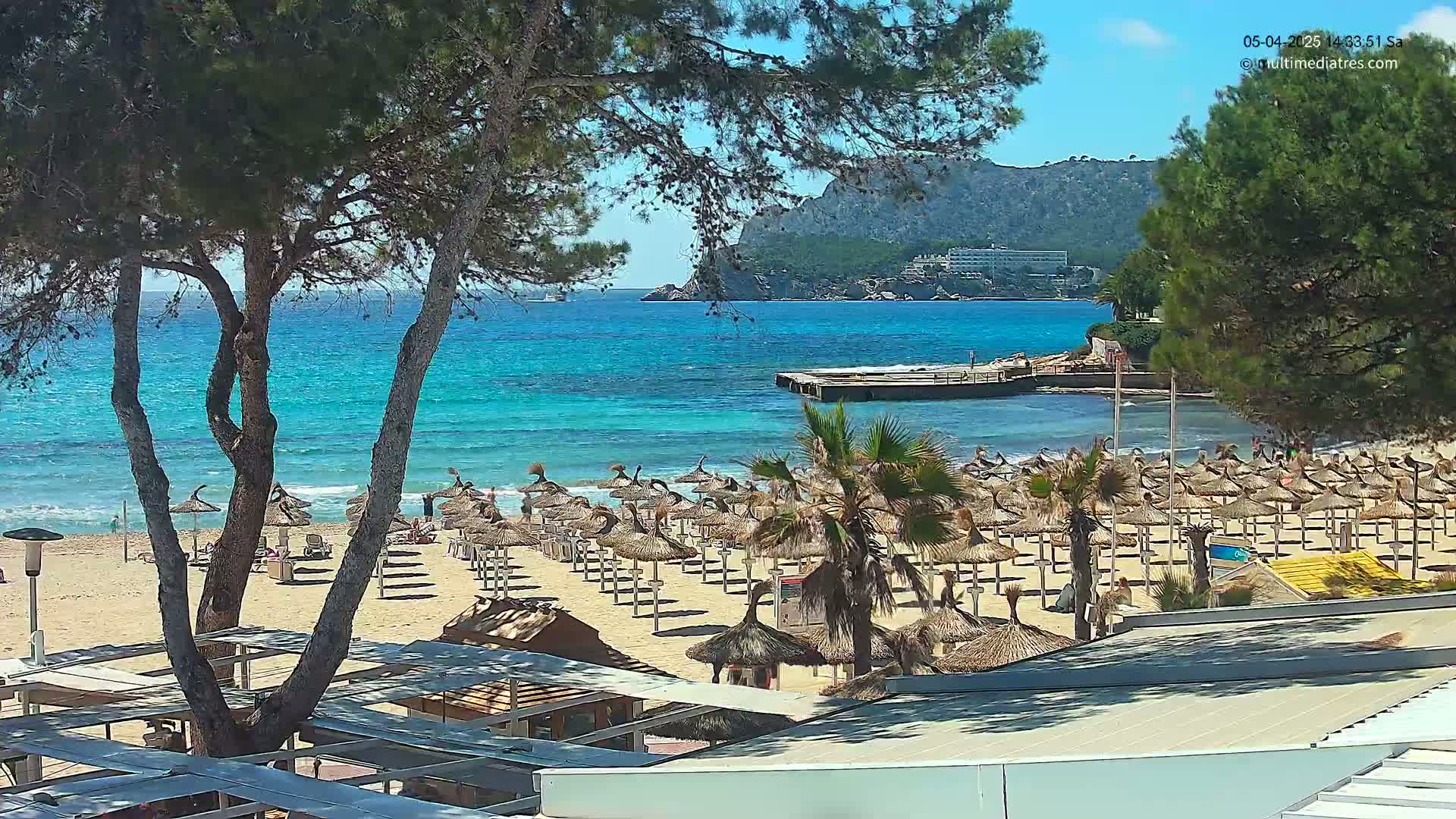 A sunny beach scene shows numerous thatched umbrellas on a sandy shore bordering a bright turquoise sea, with a rocky hillside and pine trees in the background.