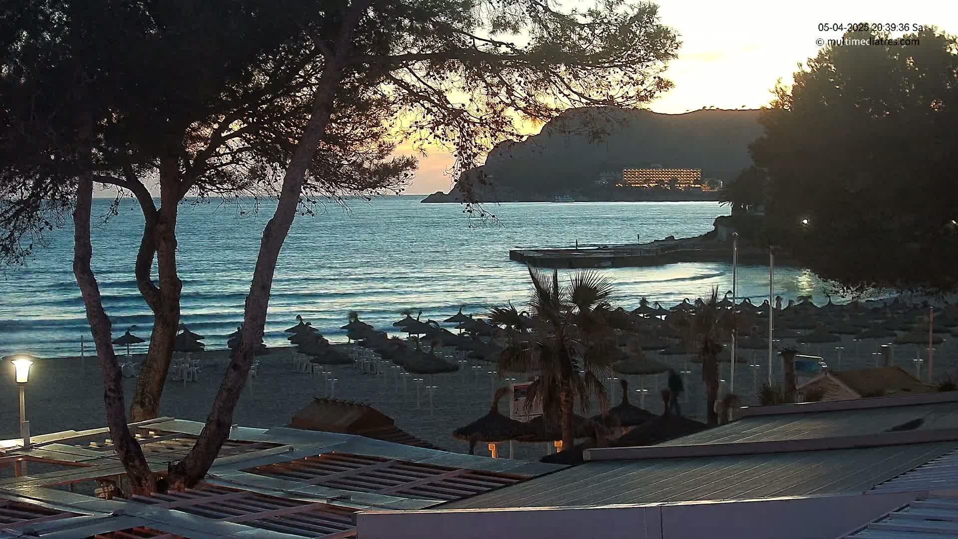 A sandy beach with numerous closed umbrellas is visible at sunset, overlooking a calm ocean with gentle waves lapping the shore, backed by a dark mountain and a distant building.
