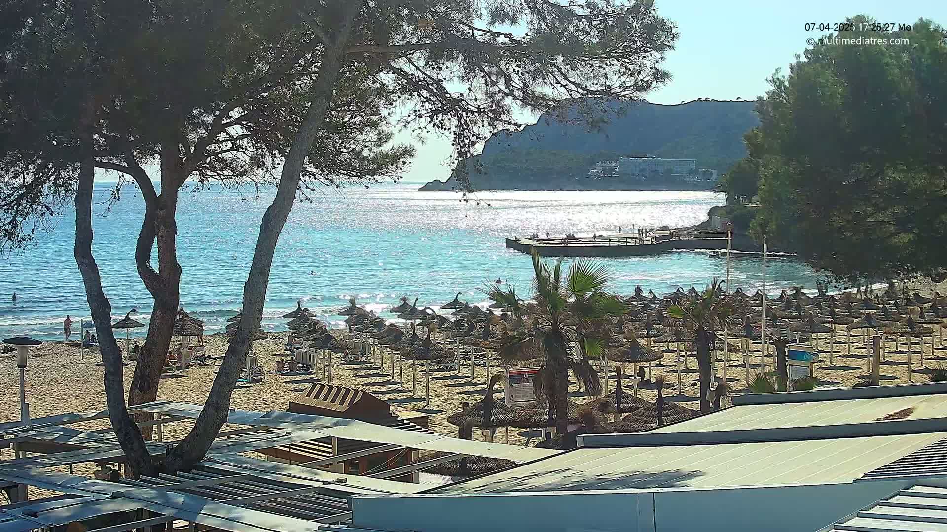 A sandy beach with numerous straw umbrellas is situated next to a calm, turquoise sea under a sunny sky, with a rocky hill visible in the background.