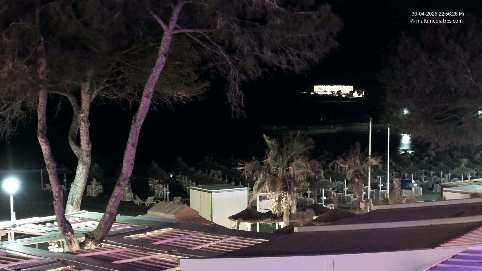 At night, a beach with rows of closed umbrellas and chairs is seen from above, with trees and a building lit up in the distance.
