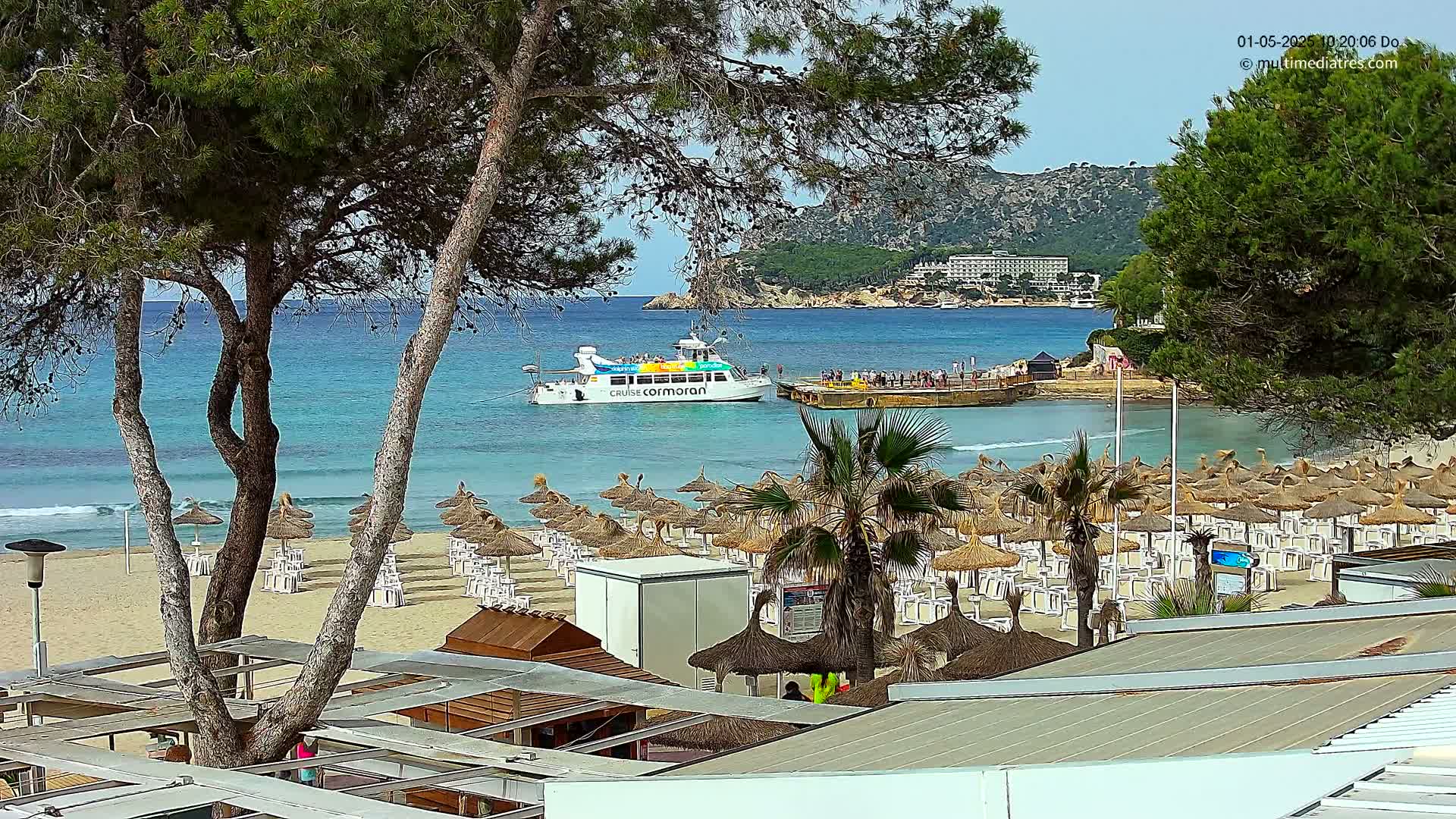 A sandy beach with numerous thatched umbrellas and lounge chairs sits beside a calm, turquoise ocean where a passenger boat is docked near a small pier;  a hilly green landscape is visible in the background.
