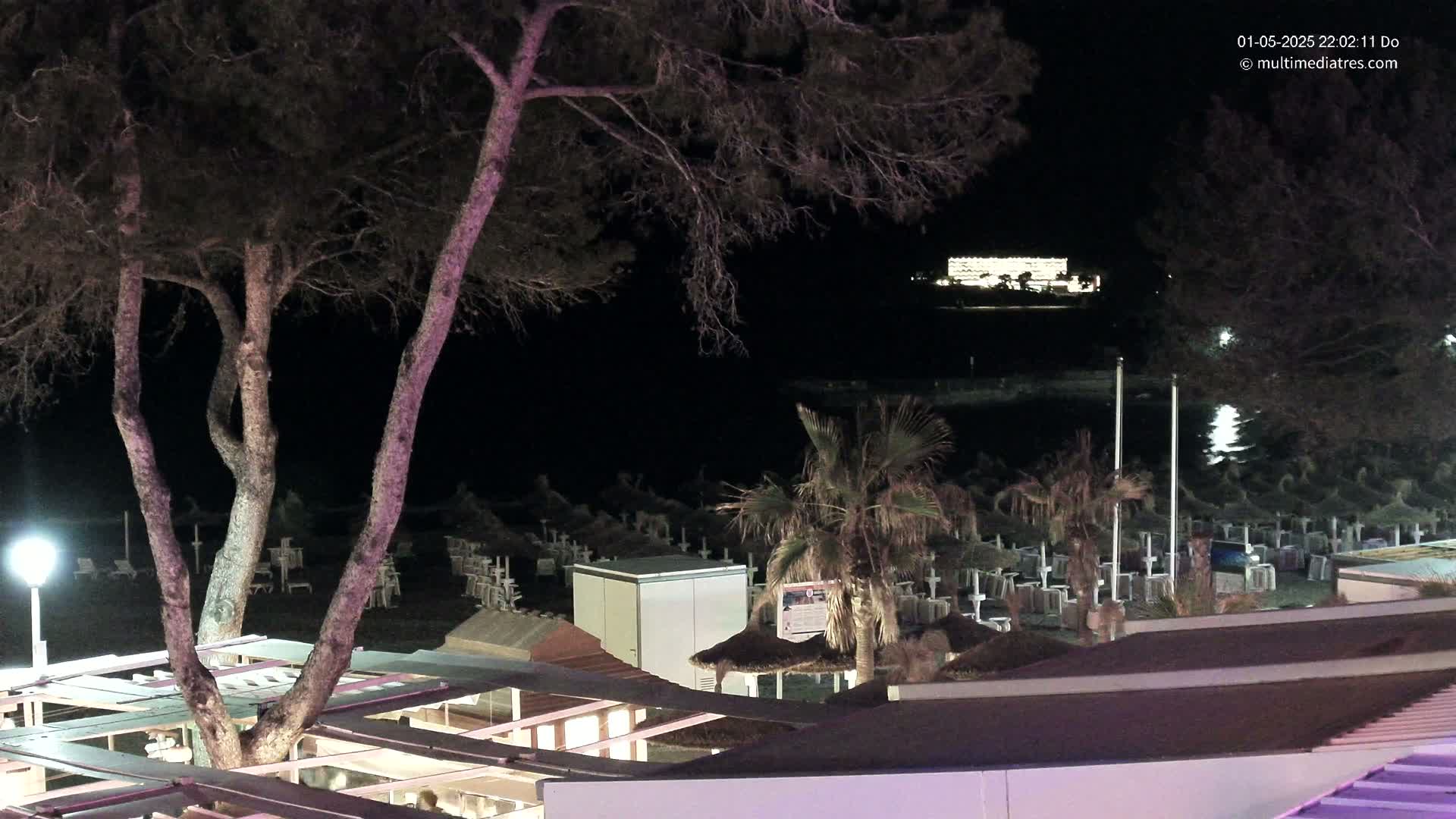 A nighttime view of a beach with closed umbrellas and beach furniture, illuminated structures, and trees, overlooking a body of water and a distant building.