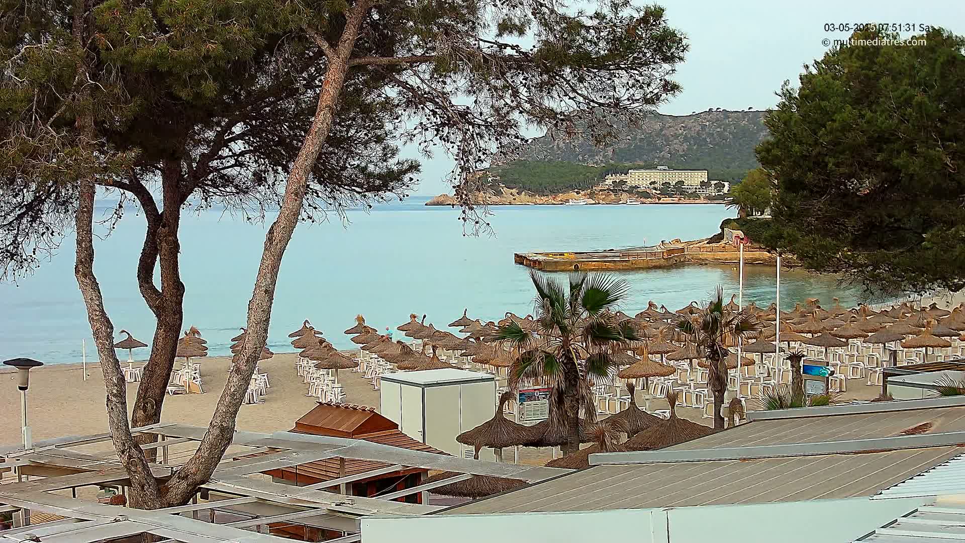 A sandy beach with numerous thatched umbrellas and chairs is seen from behind trees, overlooking a calm, light-blue sea and a distant hillside with buildings.