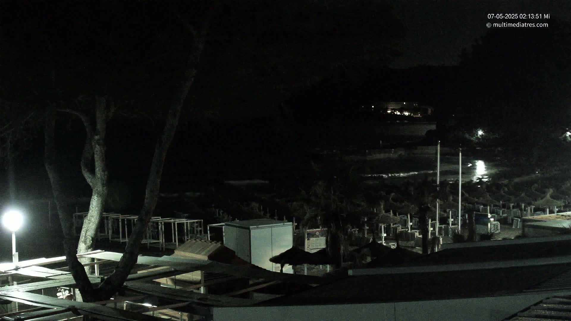A nighttime view of a beach at night shows dark trees, structures, and beach umbrellas under a mostly dark sky.