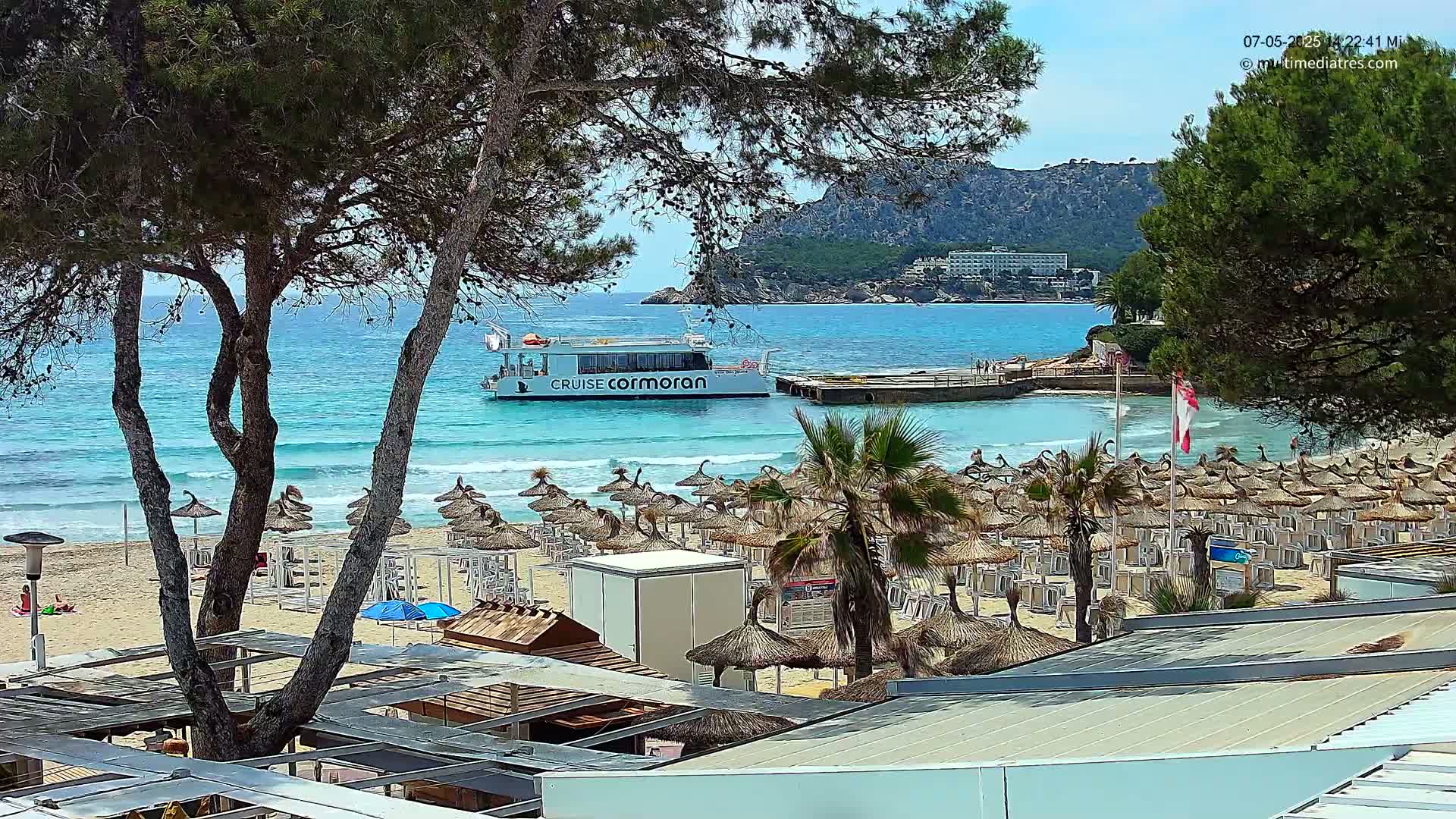 A sandy beach with numerous thatched umbrellas and lounge chairs is situated below a rocky hillside, overlooking a calm, turquoise sea where a passenger boat is docked.
