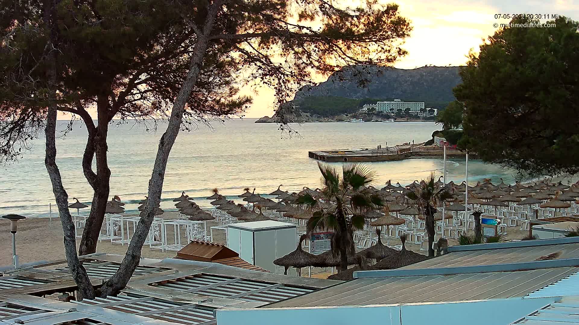 A sandy beach with numerous thatched umbrellas and white lounge chairs is situated beside calm ocean waters at sunset, with trees and a distant hotel visible in the background.