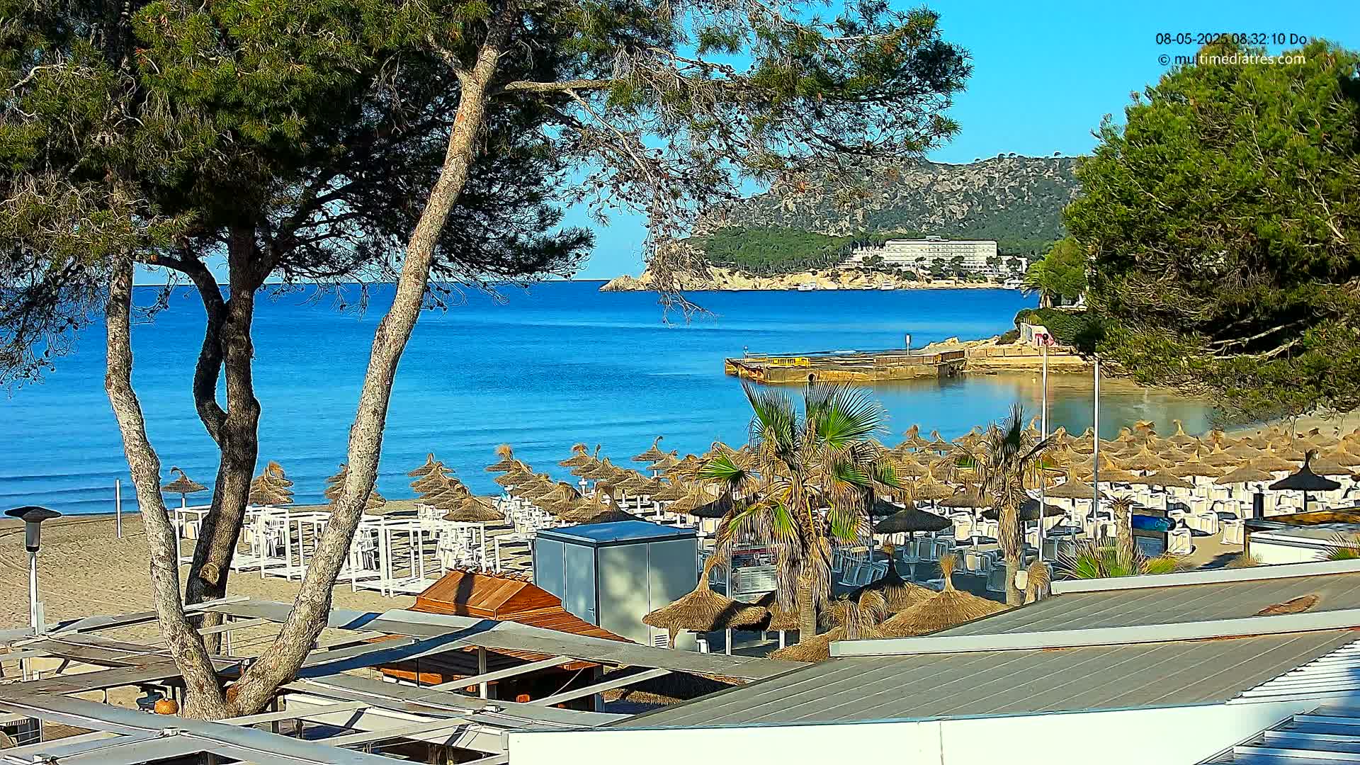 A sandy beach with numerous thatched umbrellas and white lounge chairs is situated on a calm, clear blue ocean next to a hill with lush green vegetation and a large building.