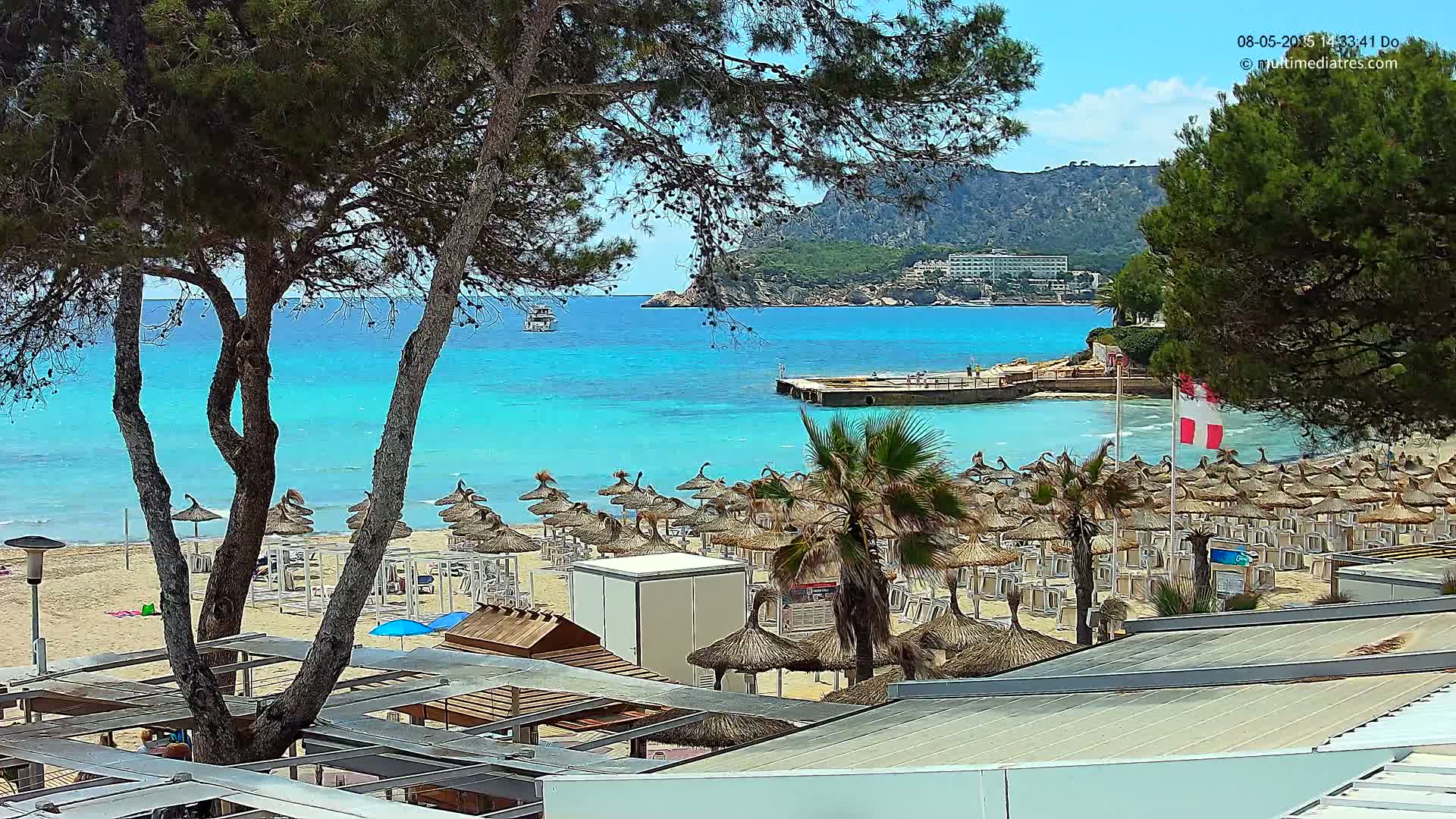 A sandy beach with numerous thatched umbrellas and lounge chairs is situated beside a vibrant turquoise sea under a sunny sky, with a distant mountain visible in the background.