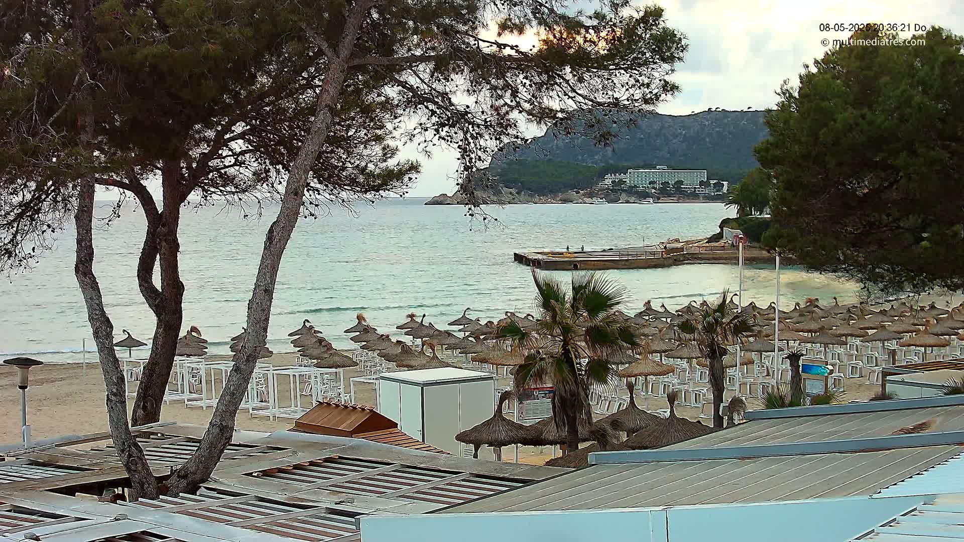 A beach with numerous thatched umbrellas and white lounge chairs sits under an overcast sky, next to a calm sea and a distant hillside with buildings.
