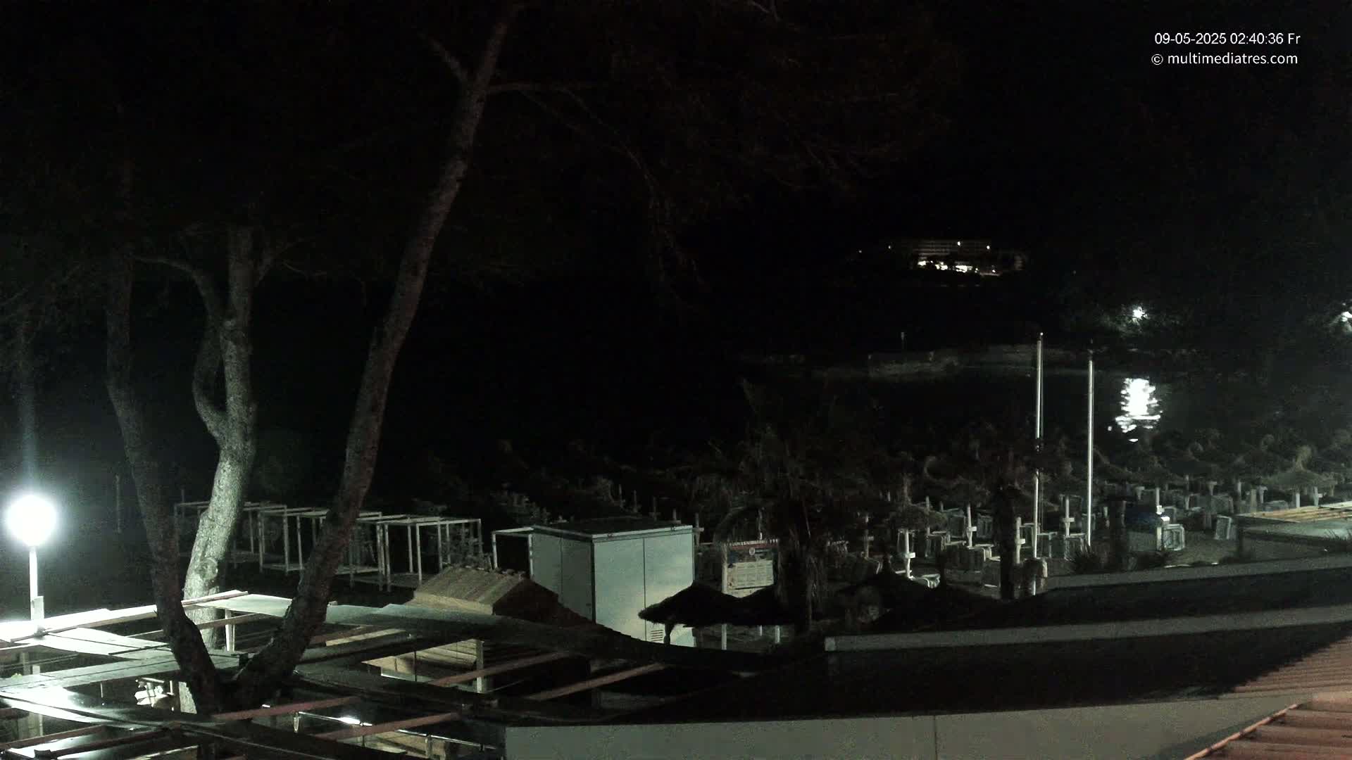 A nighttime view of a beach area with trees, structures, and what appears to be a small cemetery, illuminated by artificial light.