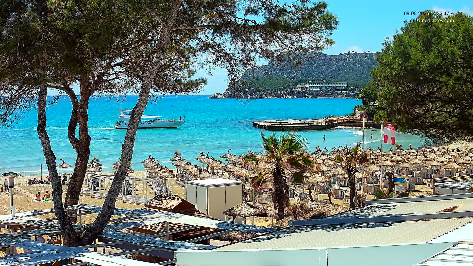 A sandy beach with numerous thatched umbrellas and lounge chairs is partially visible through the branches of pine trees, overlooking a calm, turquoise ocean where a boat is sailing toward a small dock near a rocky hillside under a sunny sky.