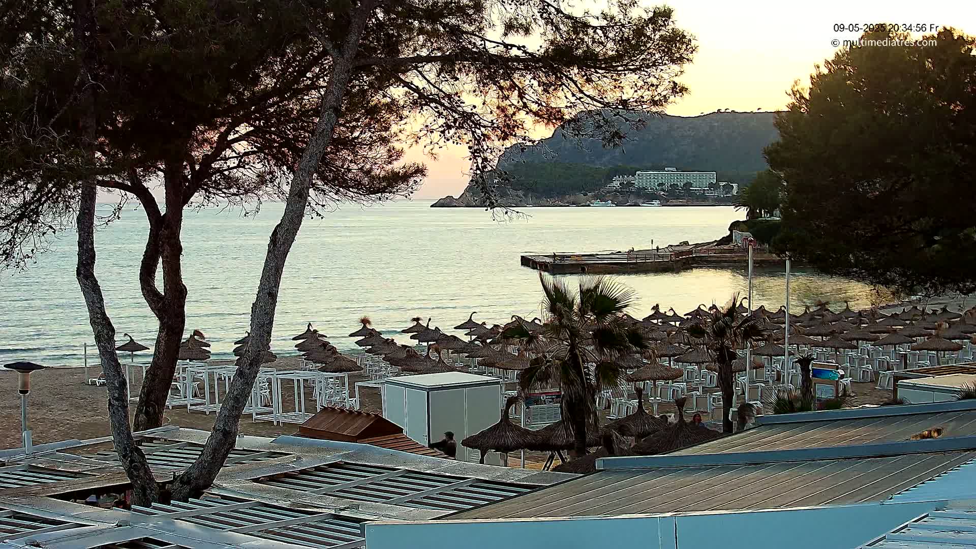 A sandy beach with numerous closed umbrellas and lounge chairs is visible under a calm sunset sky, overlooking a tranquil bay with a distant hotel and hills.