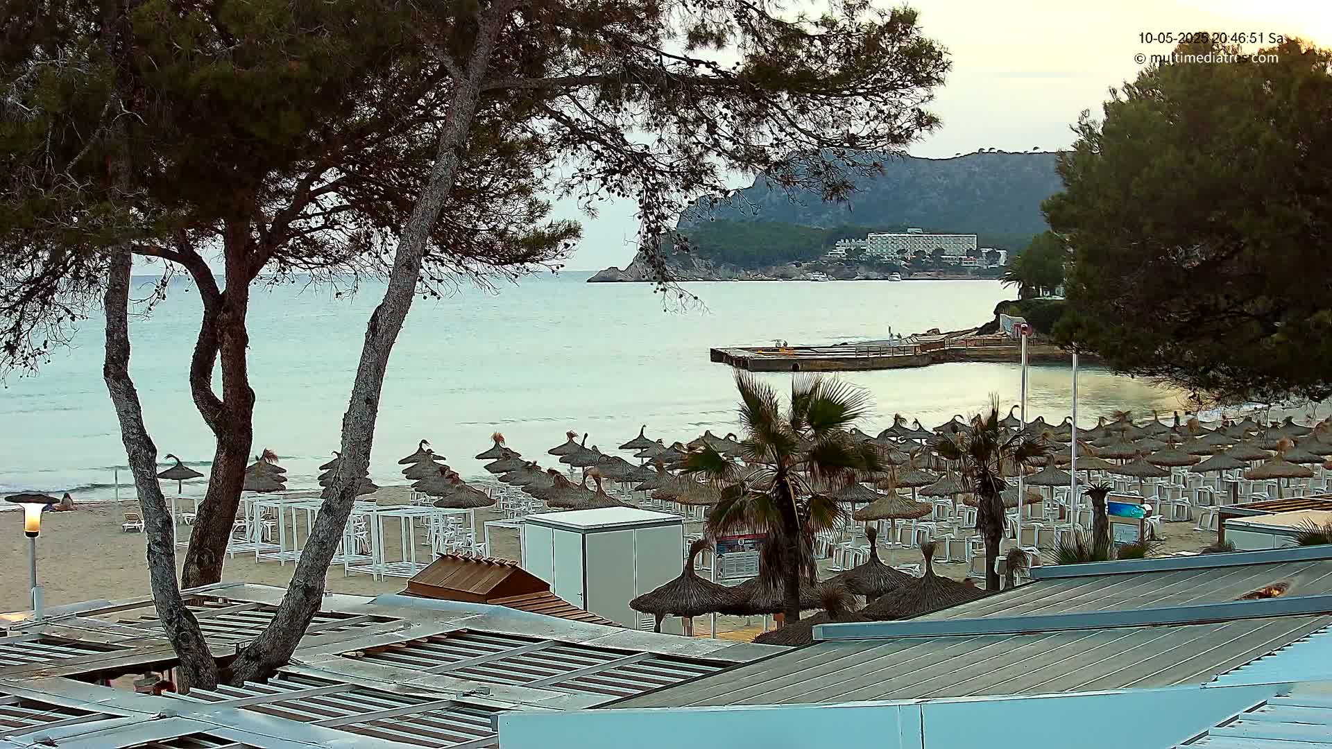 A sandy beach with numerous thatched umbrellas and lounge chairs is situated near calm, light-blue water under a partly cloudy evening sky, with a hillside visible in the background.