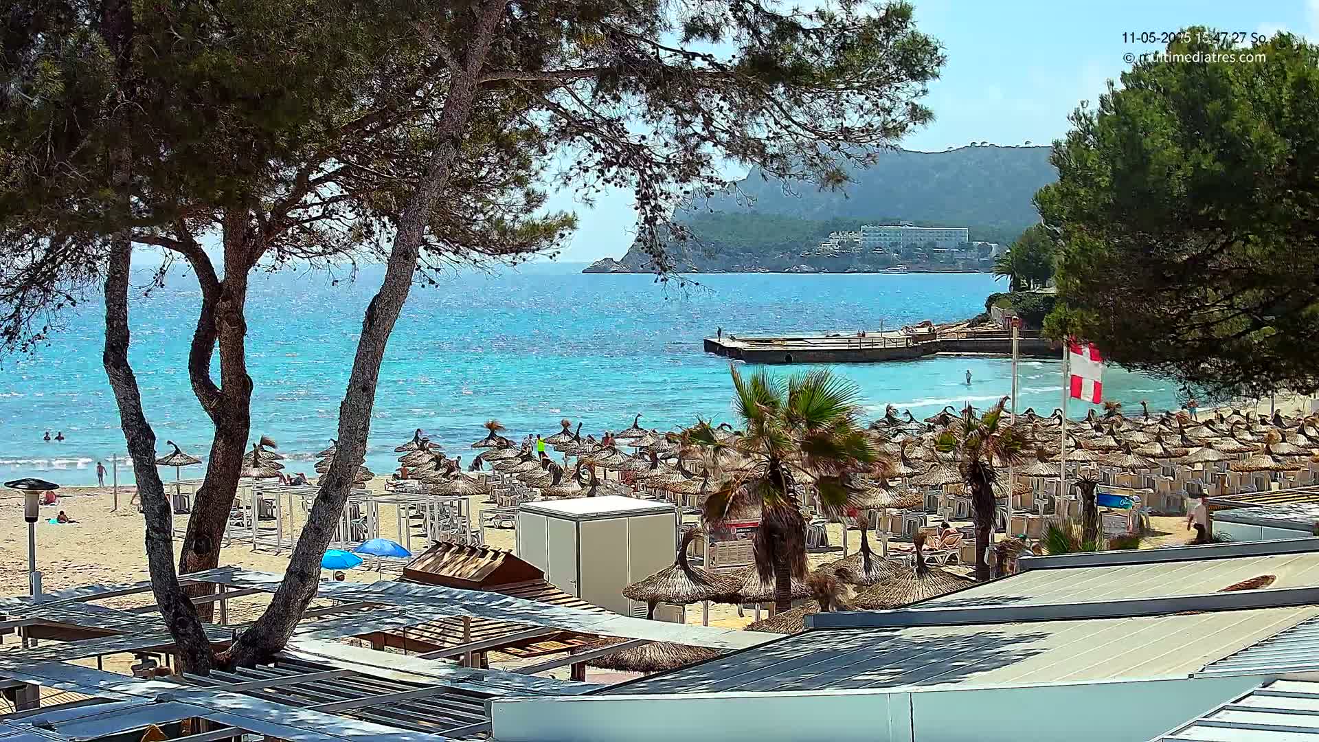 A sunny beach scene with numerous thatched umbrellas and lounge chairs on a sandy shore next to calm, turquoise water, backed by a rocky hillside and trees.