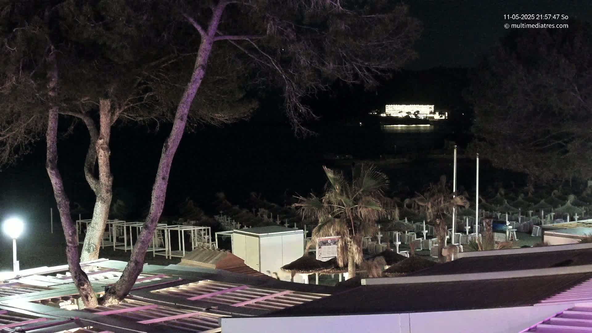 At night, a beach with closed beach umbrellas and palm trees is visible, with a brightly lit building across the water.
