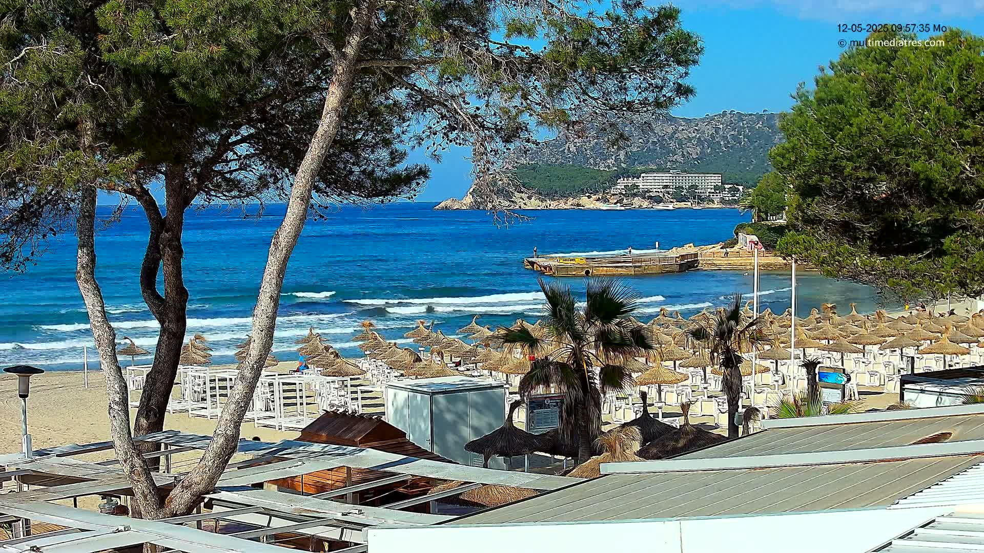 A sandy beach with numerous straw umbrellas and lounge chairs is seen under a sunny sky, overlooking a calm blue ocean with gentle waves lapping the shore, and a distant mountain range.